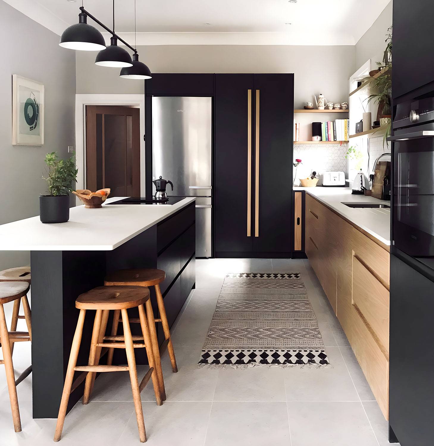 Modern kitchen featuring a wooden cabinet with a dark countertop. On the countertop, there are various ceramic bowls, plates, and a plant in a vase. A minimalist framed picture leans against a light-colored tile backsplash. A black pendant light hangs above.