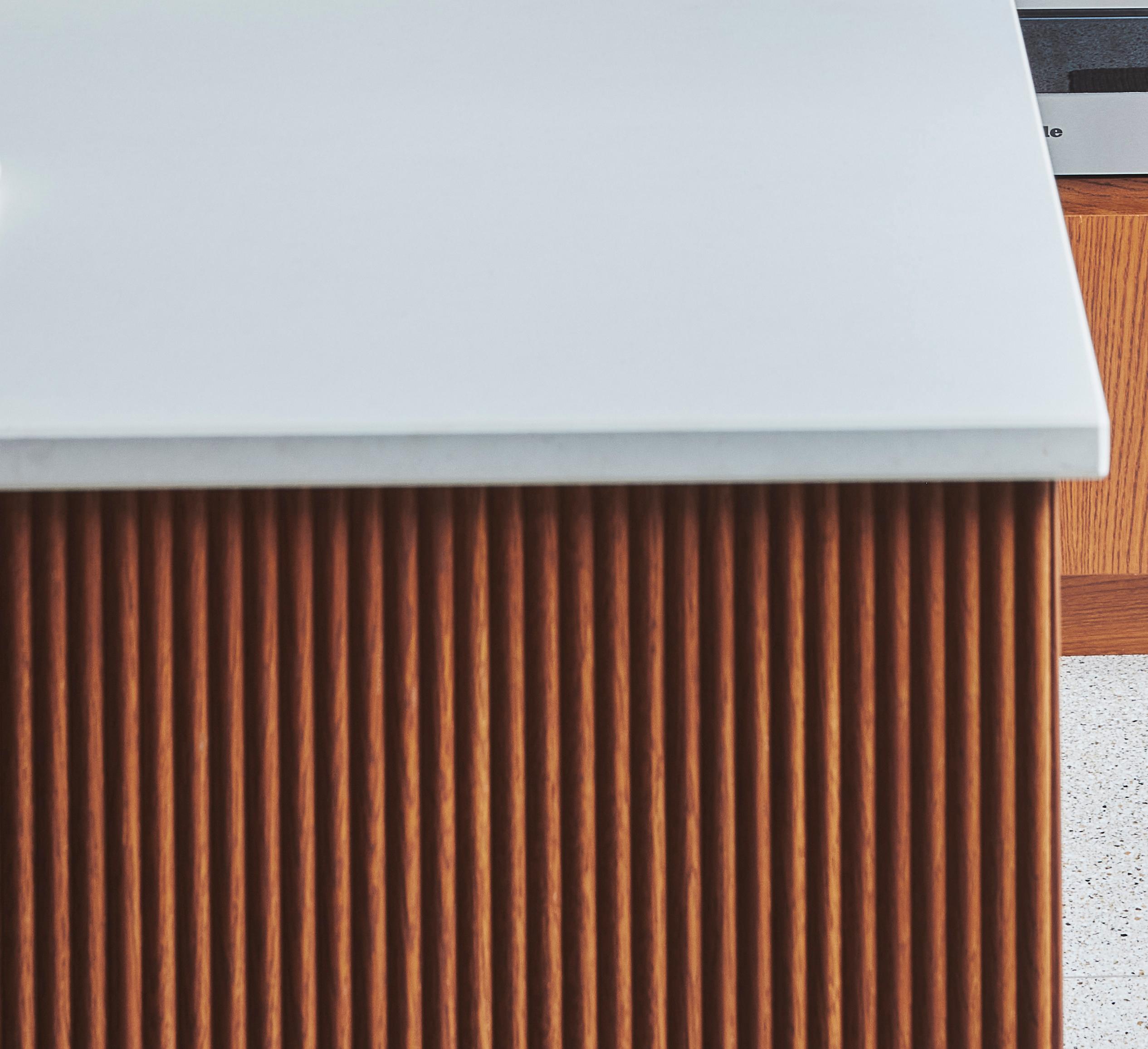 Close-up of a modern kitchen island with a white countertop. The front of the island features vertical wooden slats, creating a textured and stylish appearance. Part of a wooden cabinet and terrazzo floor can be seen in the background.