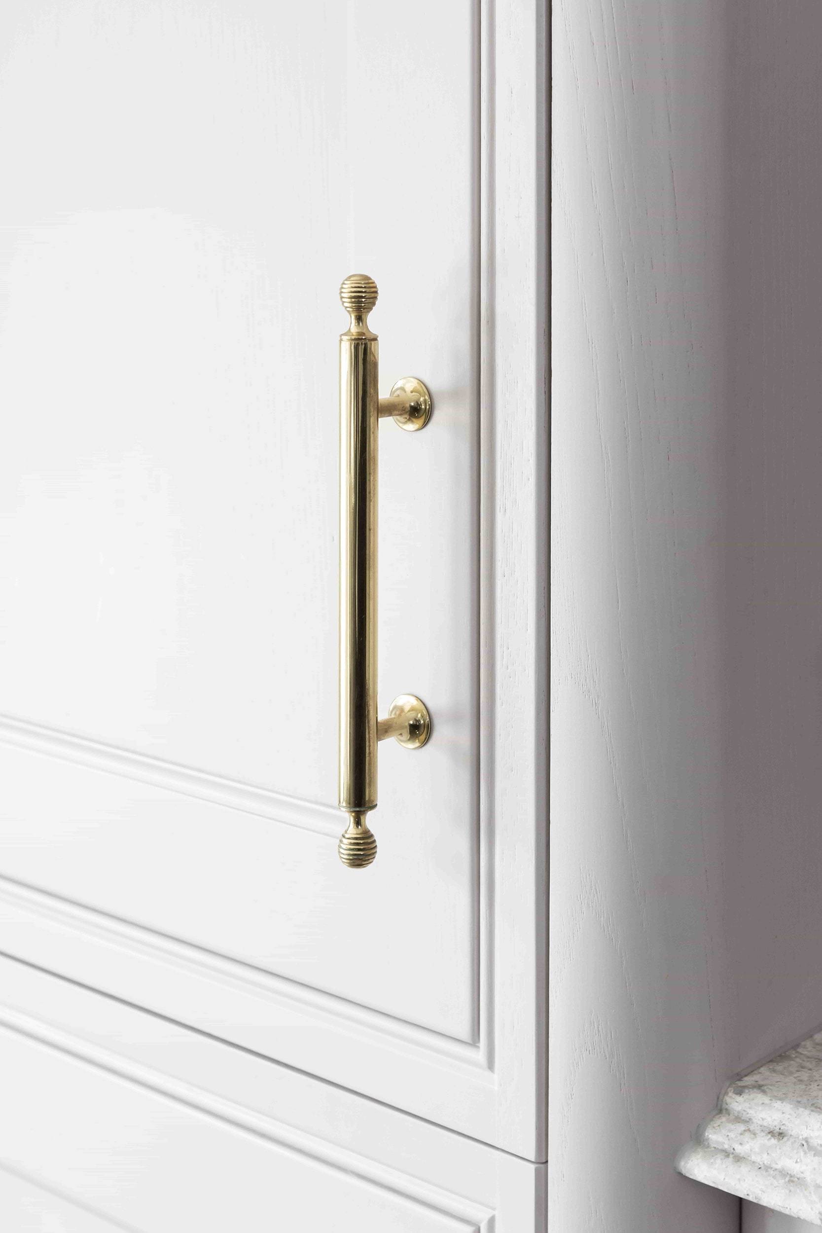 Close-up of a white cabinet door with a vertical, metallic gold handle. The handle has decorative knobs at both ends. The cabinet surface is smooth and glossy, and part of a granite countertop is visible on the right edge