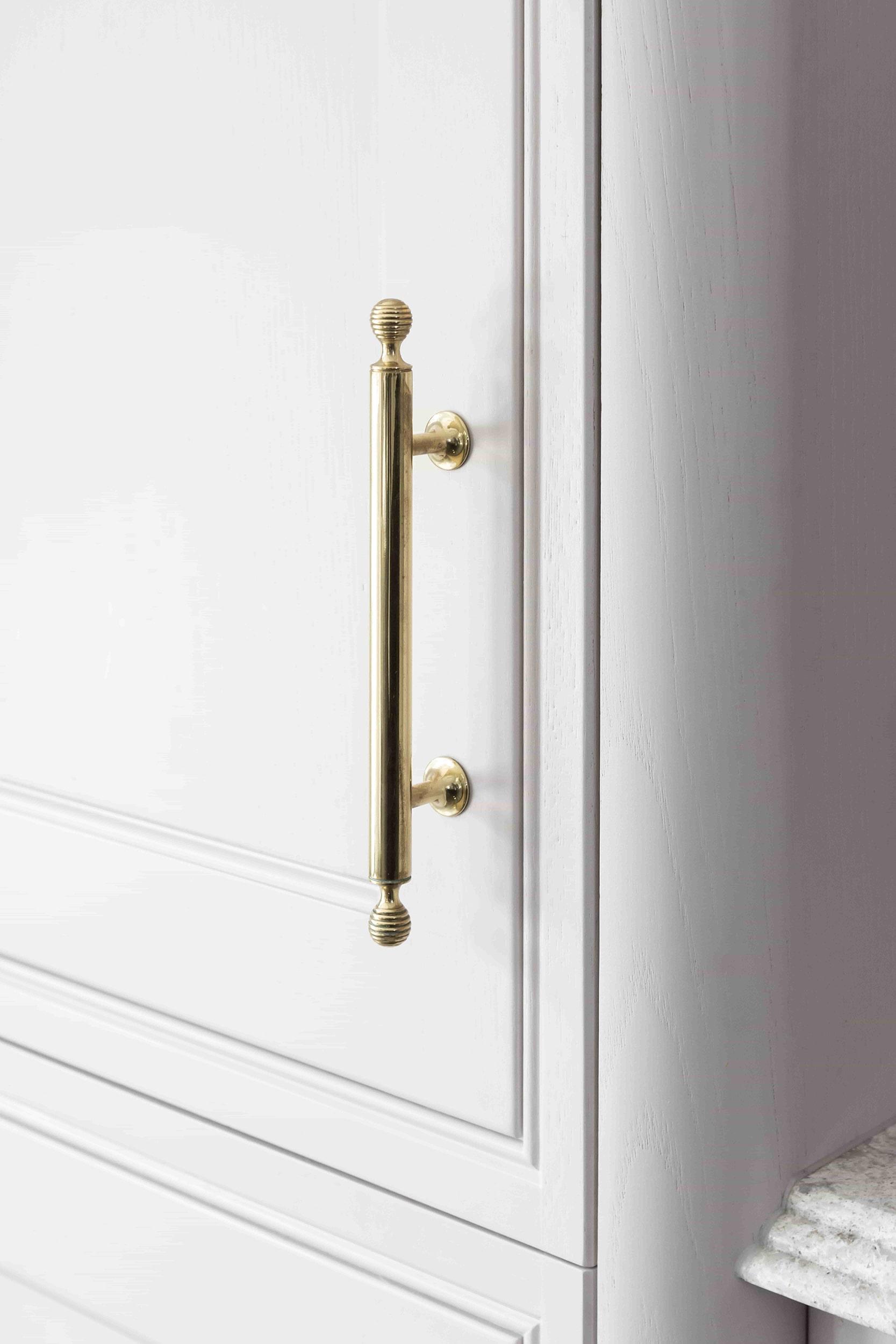 Close-up of a white cabinet door with a vertical, metallic gold handle. The handle has decorative knobs at both ends. The cabinet surface is smooth and glossy, and part of a granite countertop is visible on the right edge