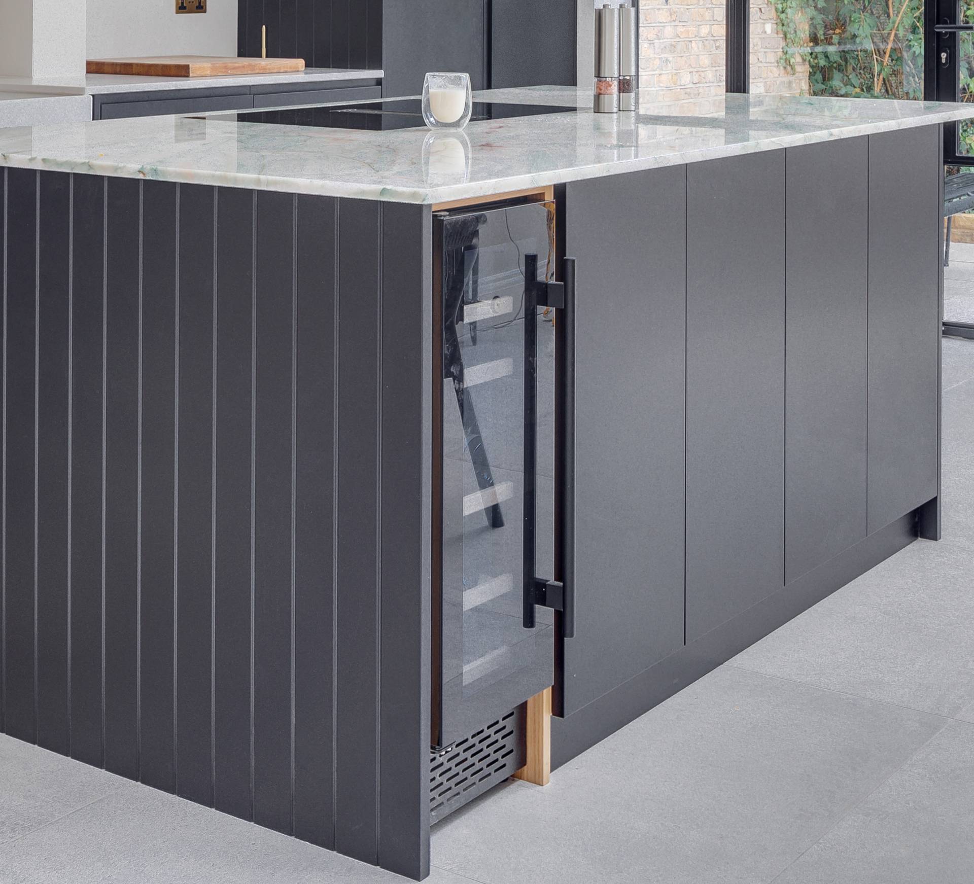 A modern kitchen island with a marble countertop features a built-in glass-front wine cooler with the door partially open, revealing shelves inside.