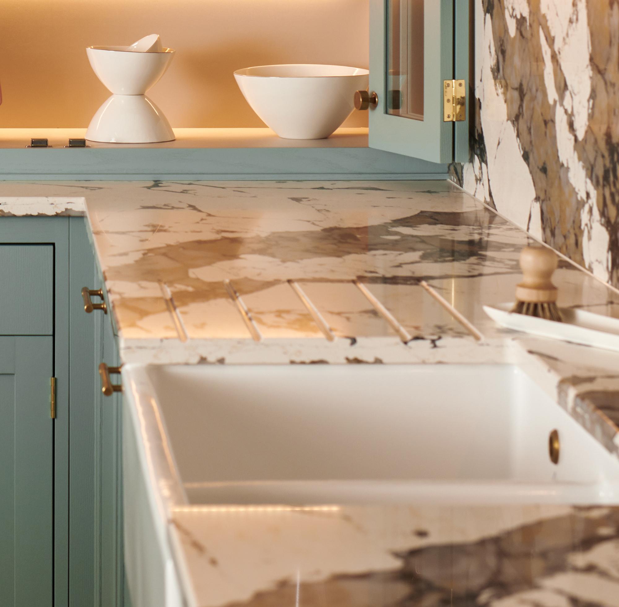 A kitchen countertop with a marble pattern featuring shades of brown and cream. It includes a white sink and teal cabinets. In the background, theres soft lighting illuminating two white bowls displayed on a shelf.