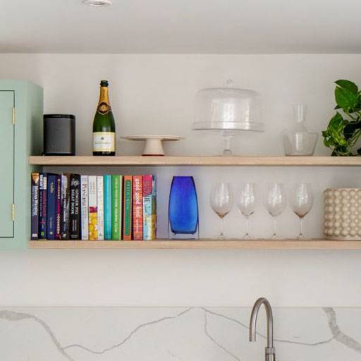 Two wooden shelves hold cookbooks, wine glasses, a blue vase, a plant, a speaker, a champagne bottle, a cake stand, and a cake dome, above a kitchen sink with a marble backsplash.