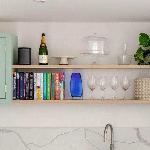 Two wooden shelves hold cookbooks, wine glasses, a blue vase, a plant, a speaker, a champagne bottle, a cake stand, and a cake dome, above a kitchen sink with a marble backsplash.