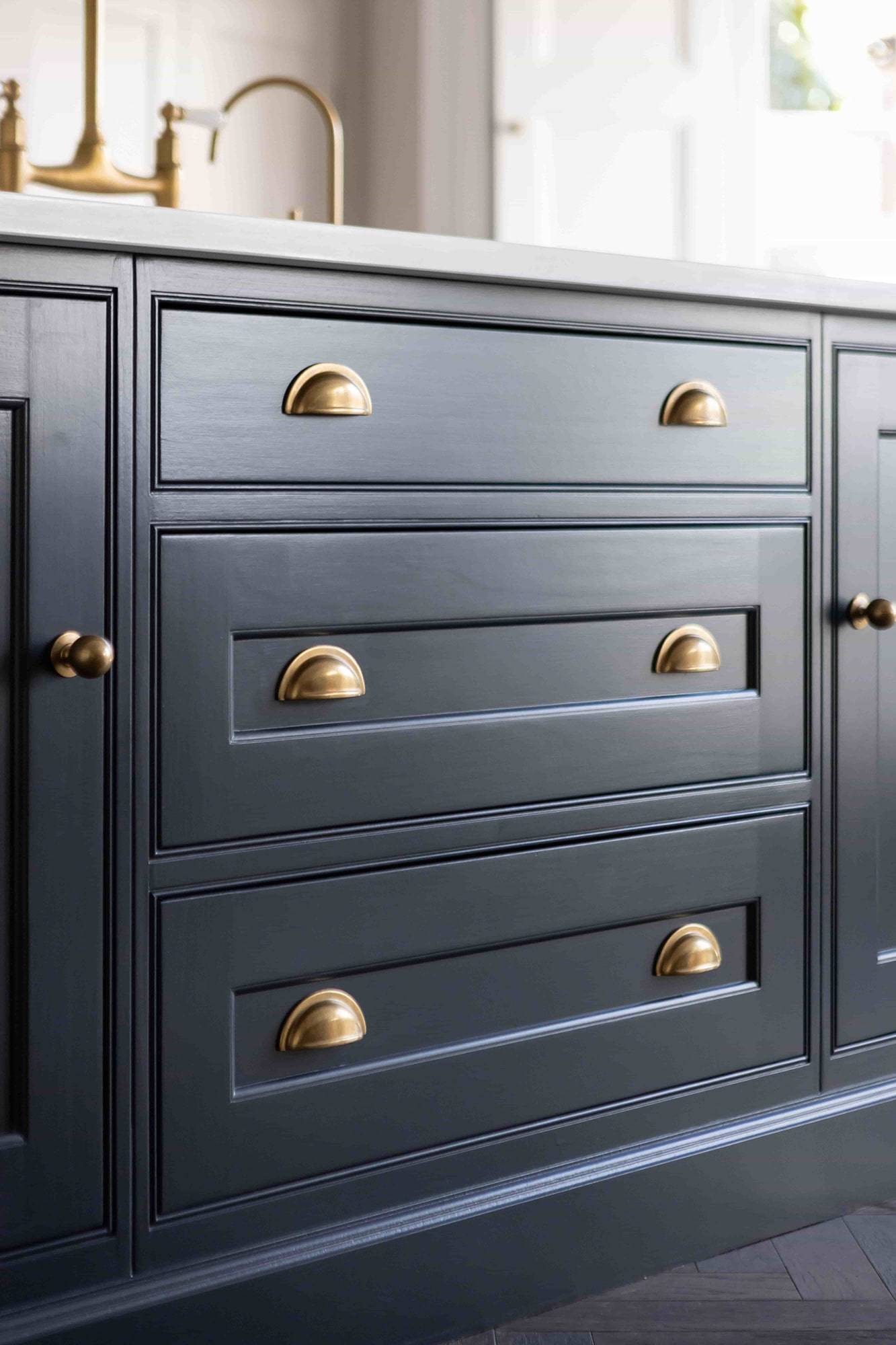 Close-up of a dark blue cabinet with three drawers and two doors. Each drawer and door features shiny brass handles, with half-cup pulls on the drawers and round knobs on the doors, creating a stylish and elegant look