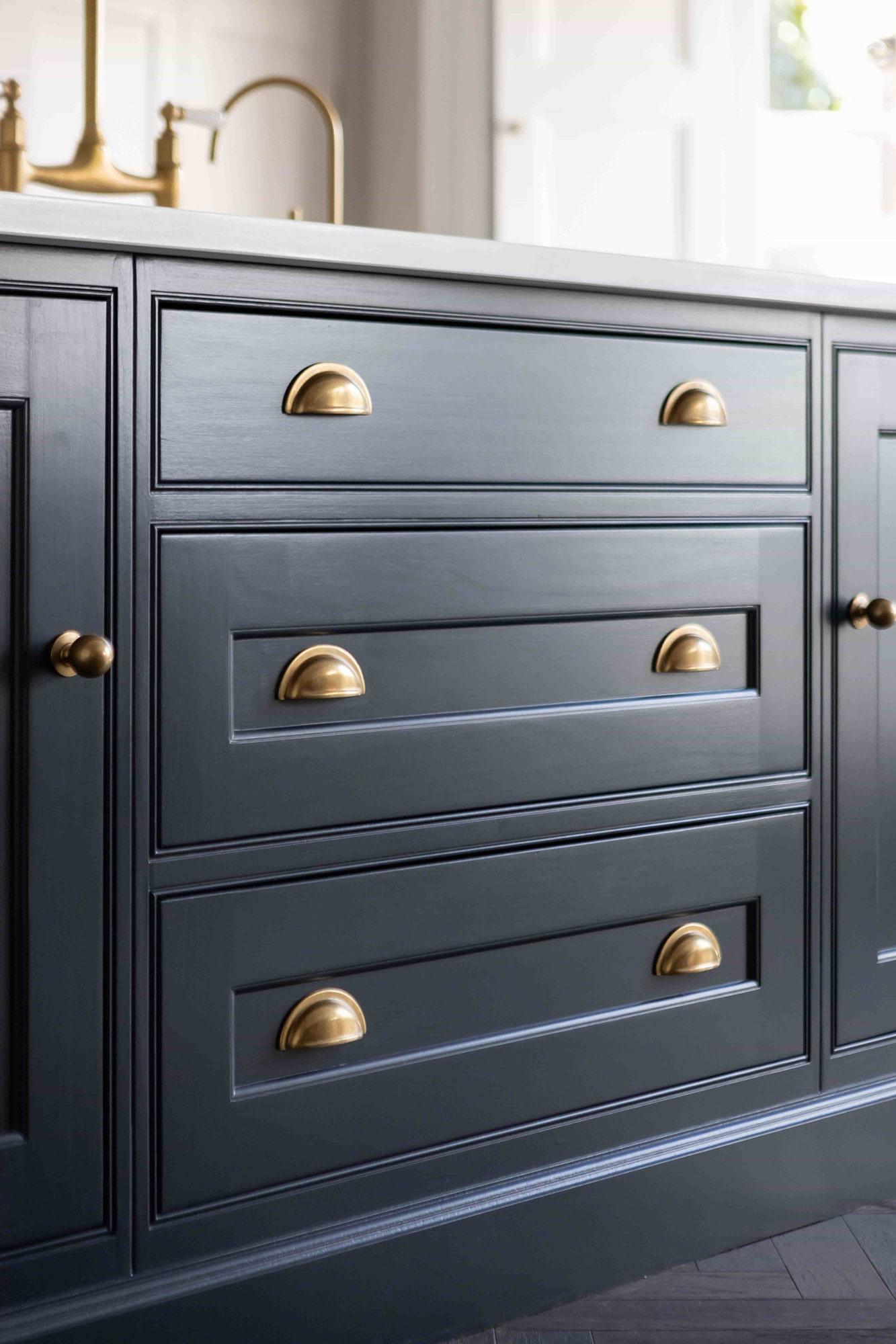 Close-up of a dark blue cabinet with three drawers and two doors. Each drawer and door features shiny brass handles, with half-cup pulls on the drawers and round knobs on the doors, creating a stylish and elegant look