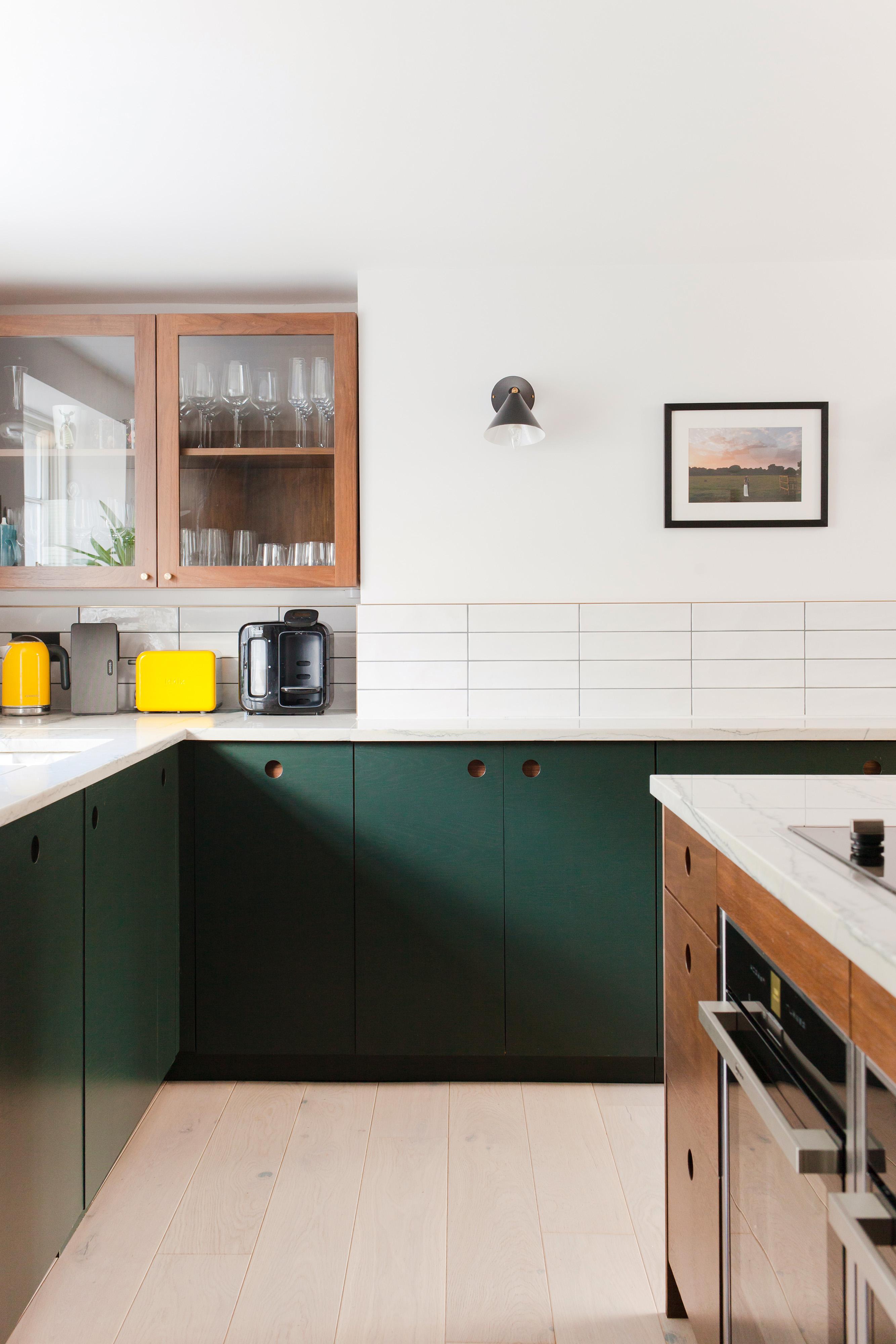 Modern kitchen with green cabinets and white countertops. Includes a wall-mounted lamp, a small framed picture, wooden shelves with glassware, and appliances like a toaster and coffee maker. Light wood flooring complements the clean design