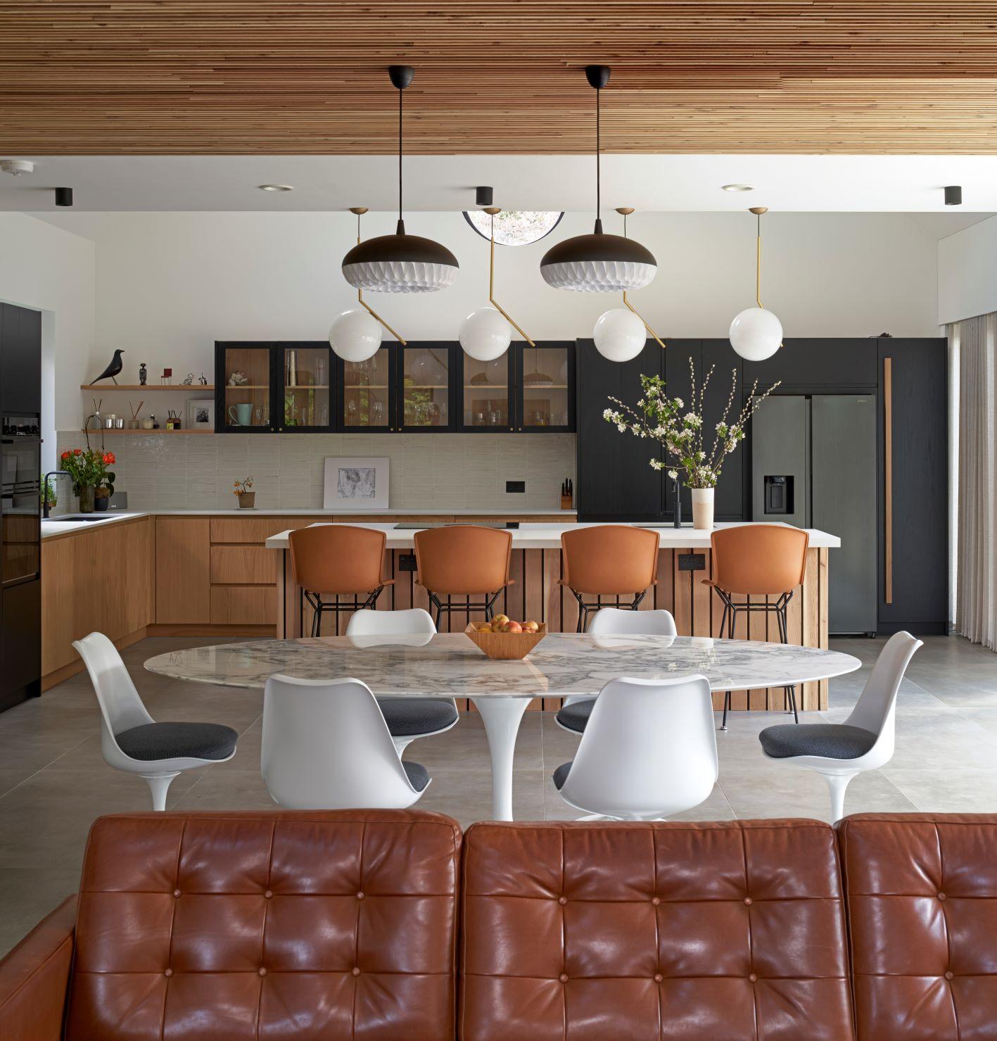 A modern kitchen and dining area with a wooden ceiling, featuring a marble table surrounded by white chairs, a brown leather couch in the foreground, and an island with brown stools. Pendant lights hang above the island.
