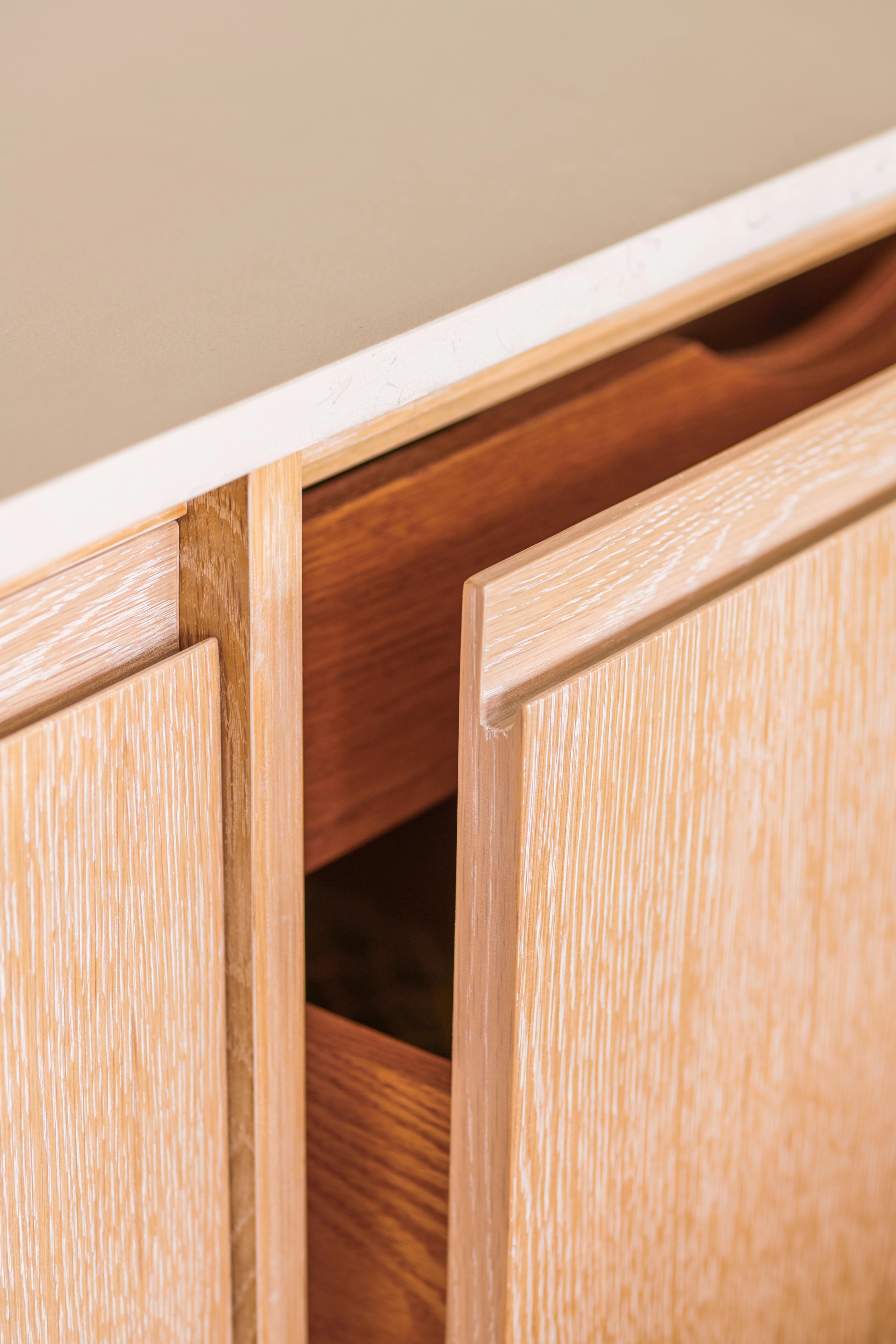 a close-up shot highlights a light-colored, textured wooden kitchen cabinet door slightly open to reveal a darker wooden interior drawer underneath a light beige countertop.