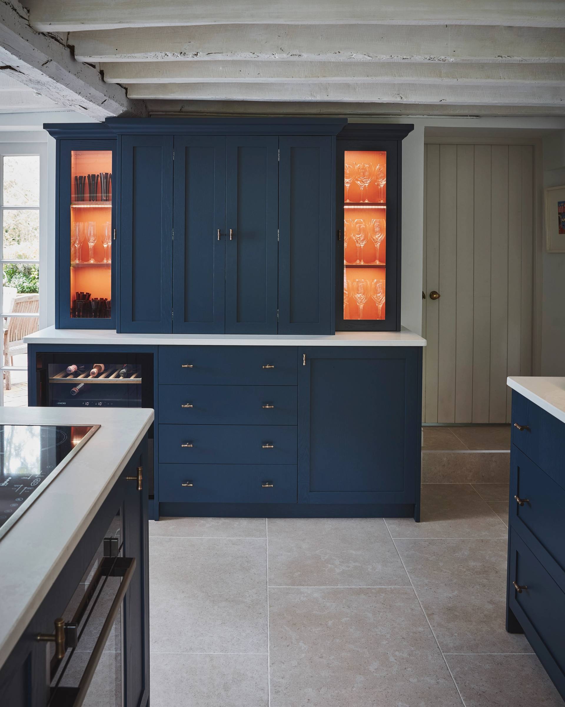 A modern kitchen with navy blue cabinets, white countertops, and glass display shelves illuminated with orange lighting, showcasing glassware. The floor is tiled, and exposed white ceiling beams are visible.