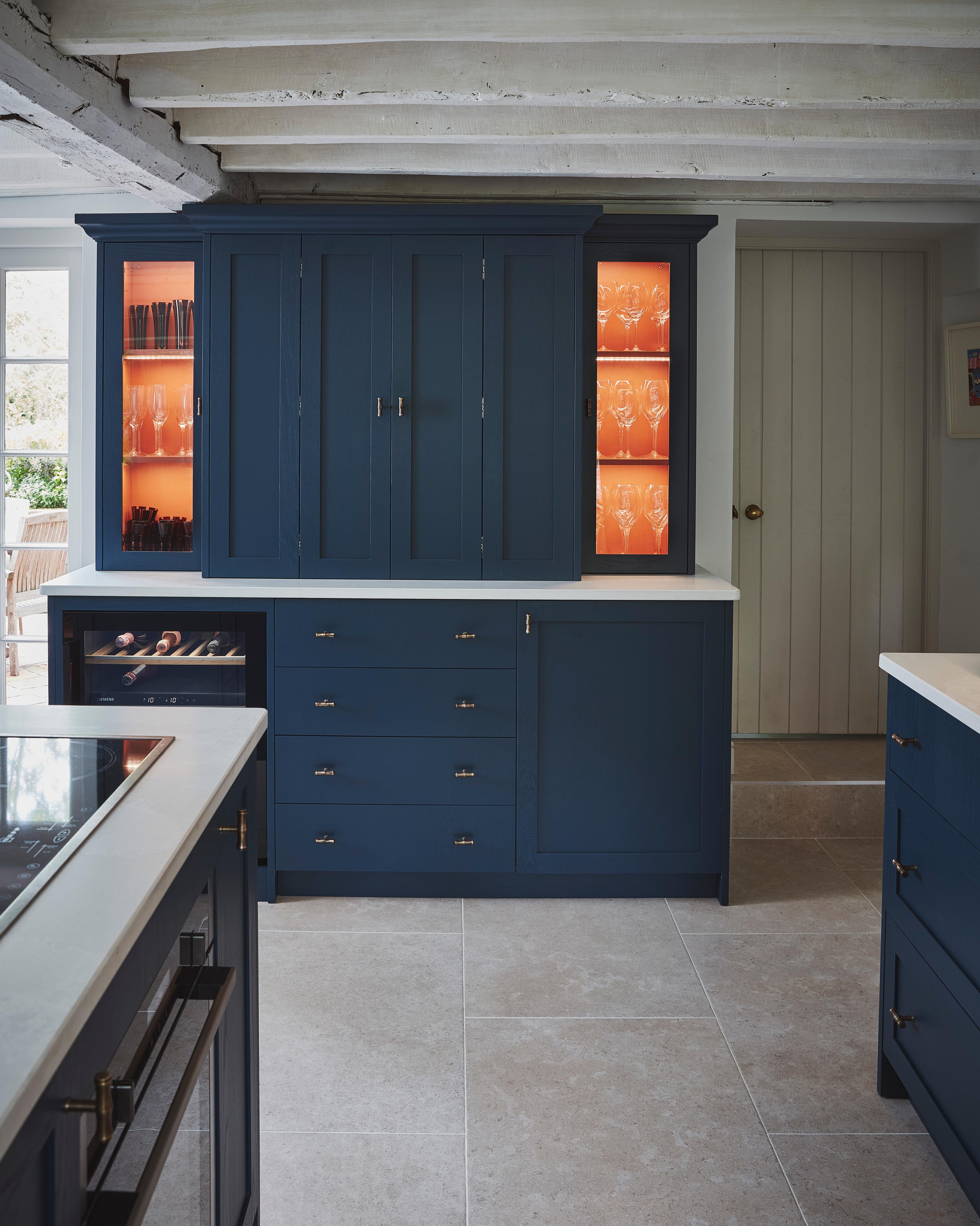A modern kitchen with navy blue cabinets, white countertops, and glass display shelves illuminated with orange lighting, showcasing glassware. The floor is tiled, and exposed white ceiling beams are visible.