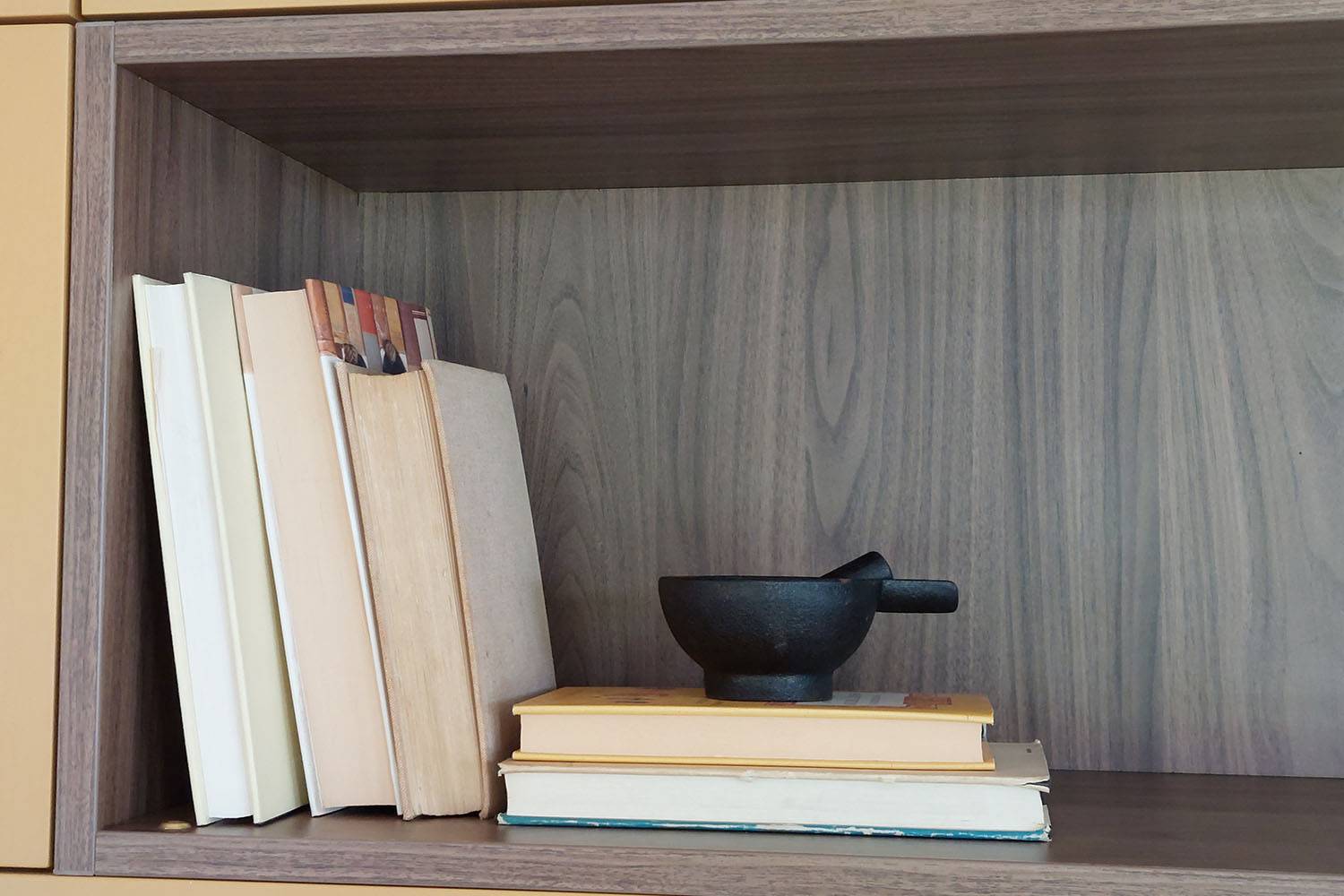 A wooden shelf with a row of books leaning against the left side. In front of the books, there are three additional books stacked horizontally. A small black mortar is placed on the stacked books.