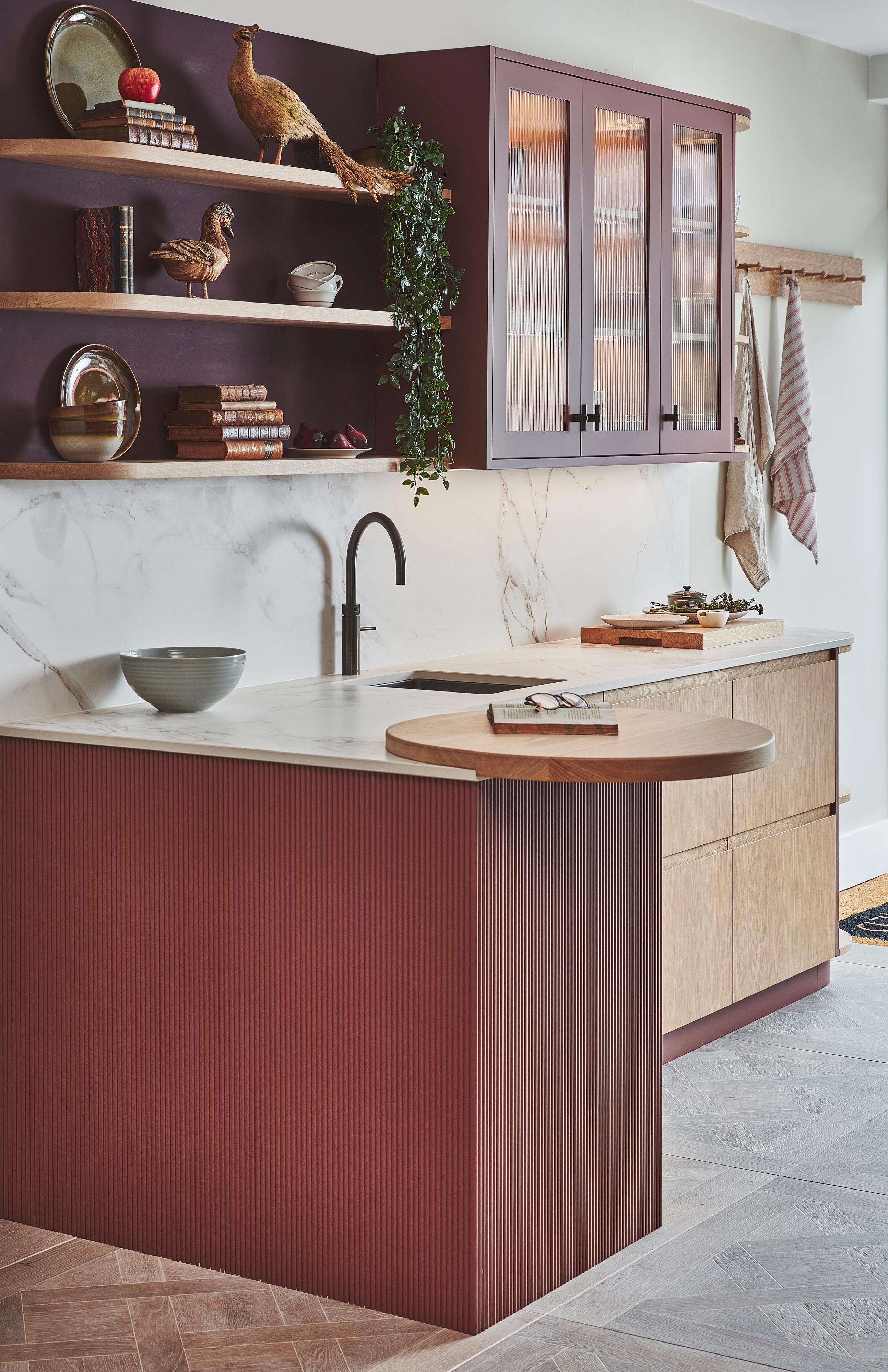 Modern kitchen with burgundy and wood tones. Open shelves display books, plates, and decorative birds. Marble backsplash and countertop enhance the design. A green plant hangs over the counter, and there are towels next to the stove area