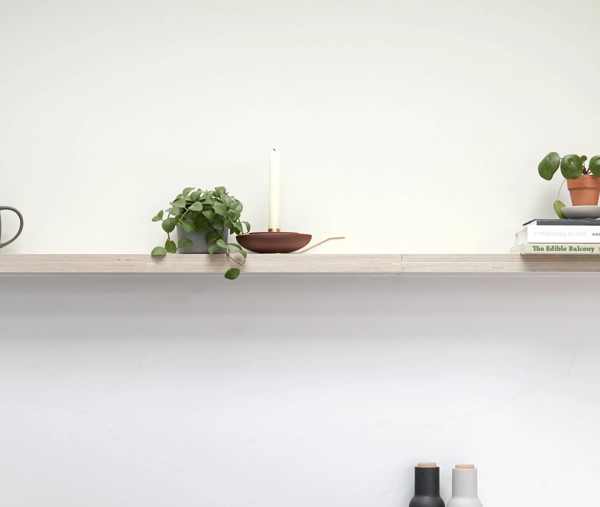 A minimal wooden shelf with a potted plant, a candle in a holder, and stacked books with another small plant on top. The background is plain white, giving a clean and simple look.