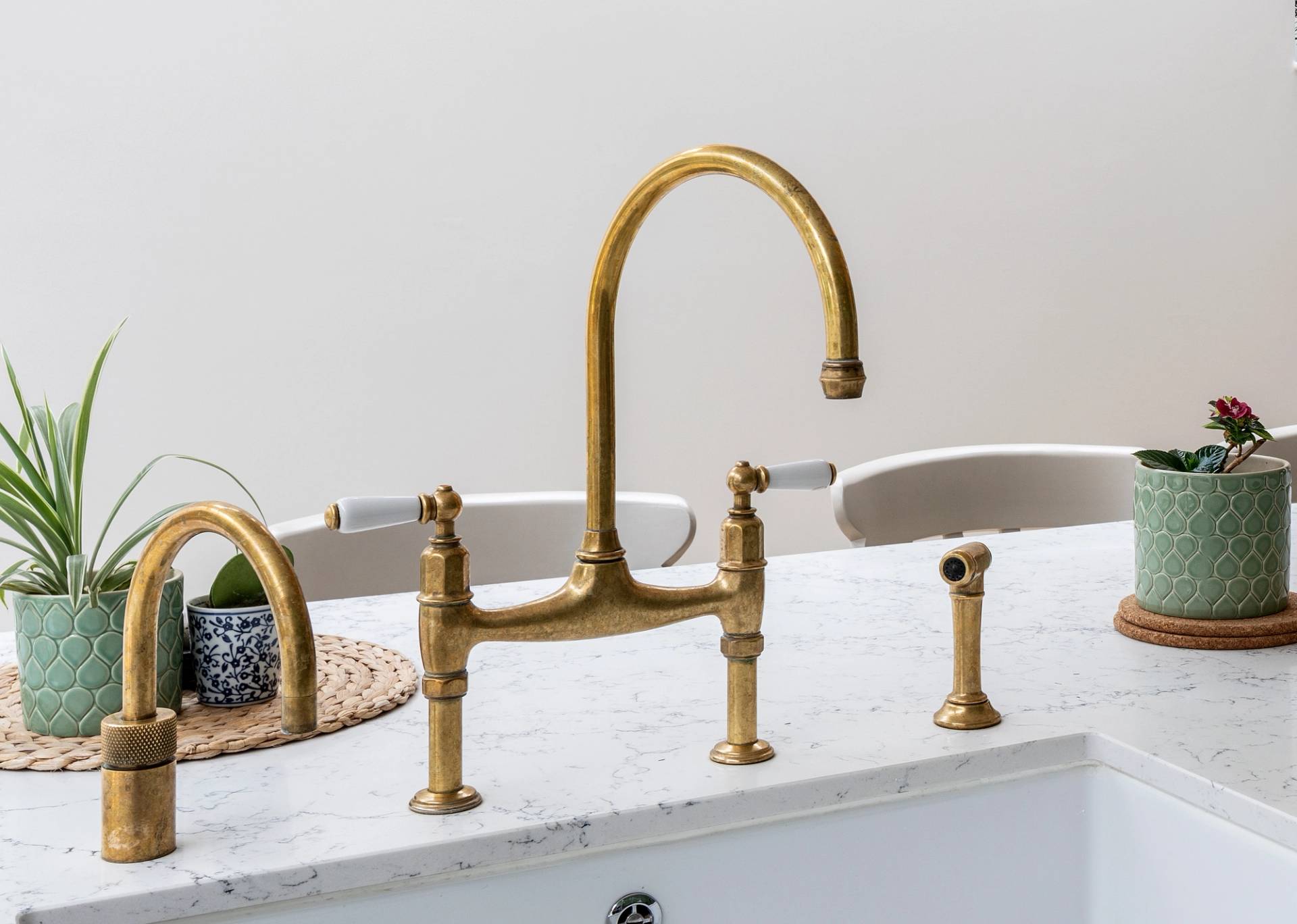 A brass kitchen faucet with a high curved spout on a white marble countertop, surrounded by green potted plants and beige chairs in the background