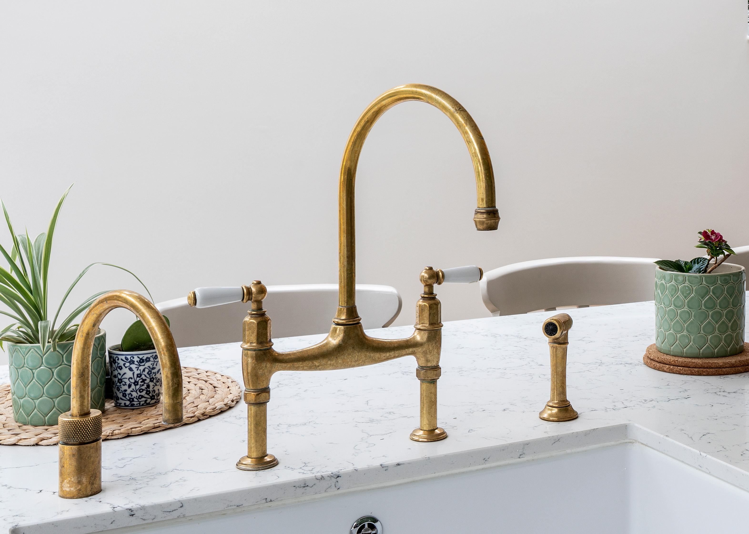 A brass kitchen faucet with a high curved spout on a white marble countertop, surrounded by green potted plants and beige chairs in the background