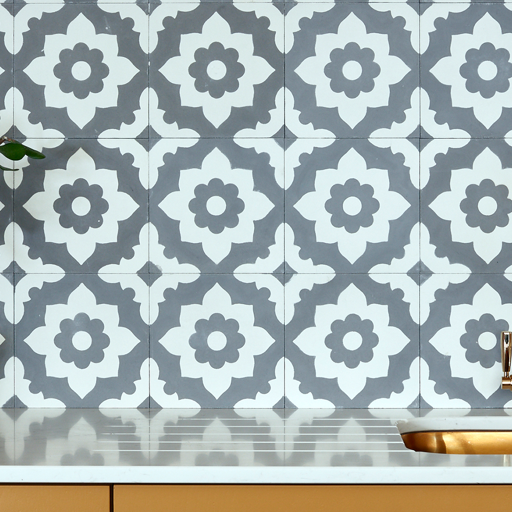 A backsplash with gray and white geometric floral tiles above a white countertop. A gold-toned sink and faucet are partially visible, and a small green plant is on the left.
