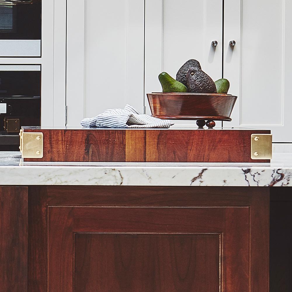 A wooden kitchen island with a marble countertop, topped with a large wooden cutting board, a folded striped towel, and a copper fruit bowl holding avocados and pears. White cabinets are in the background.