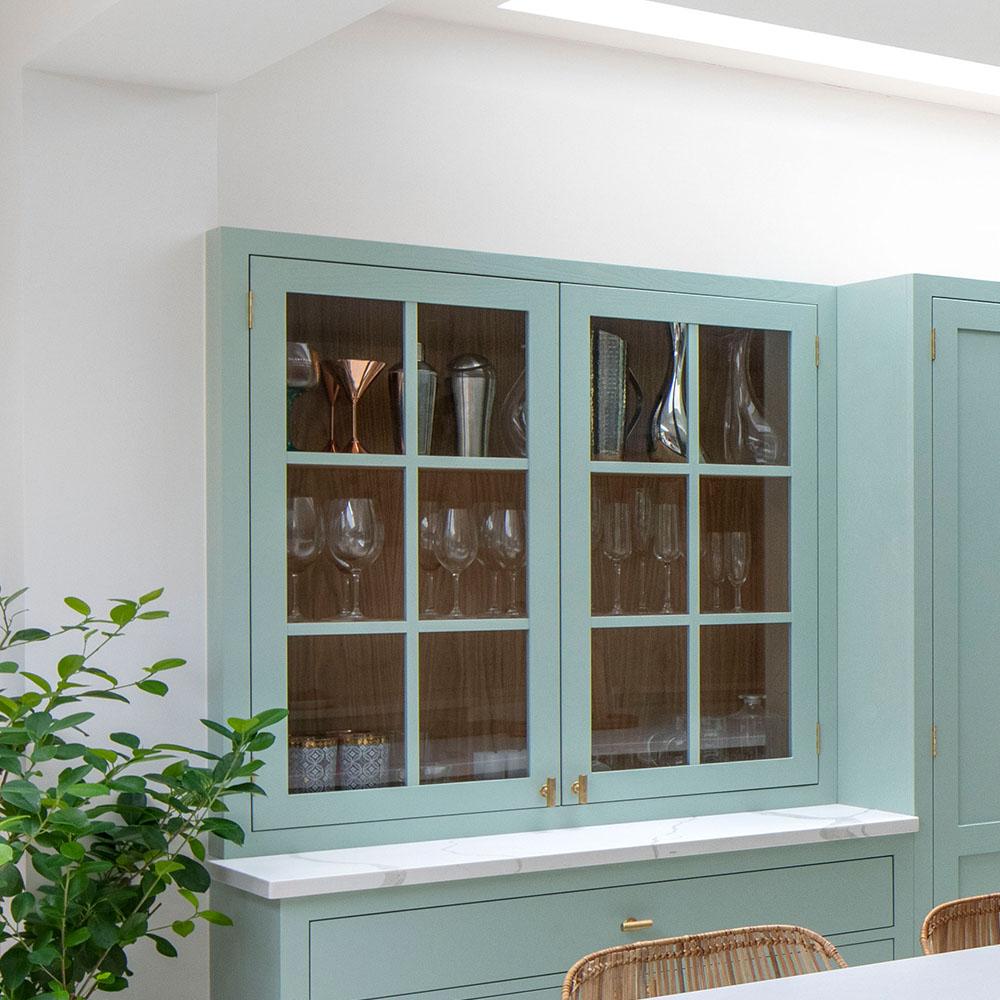 Light blue-green kitchen cabinets with glass doors display assorted glassware, including wine glasses and vases. A marble countertop and a leafy green plant are also visible in the bright, modern space.