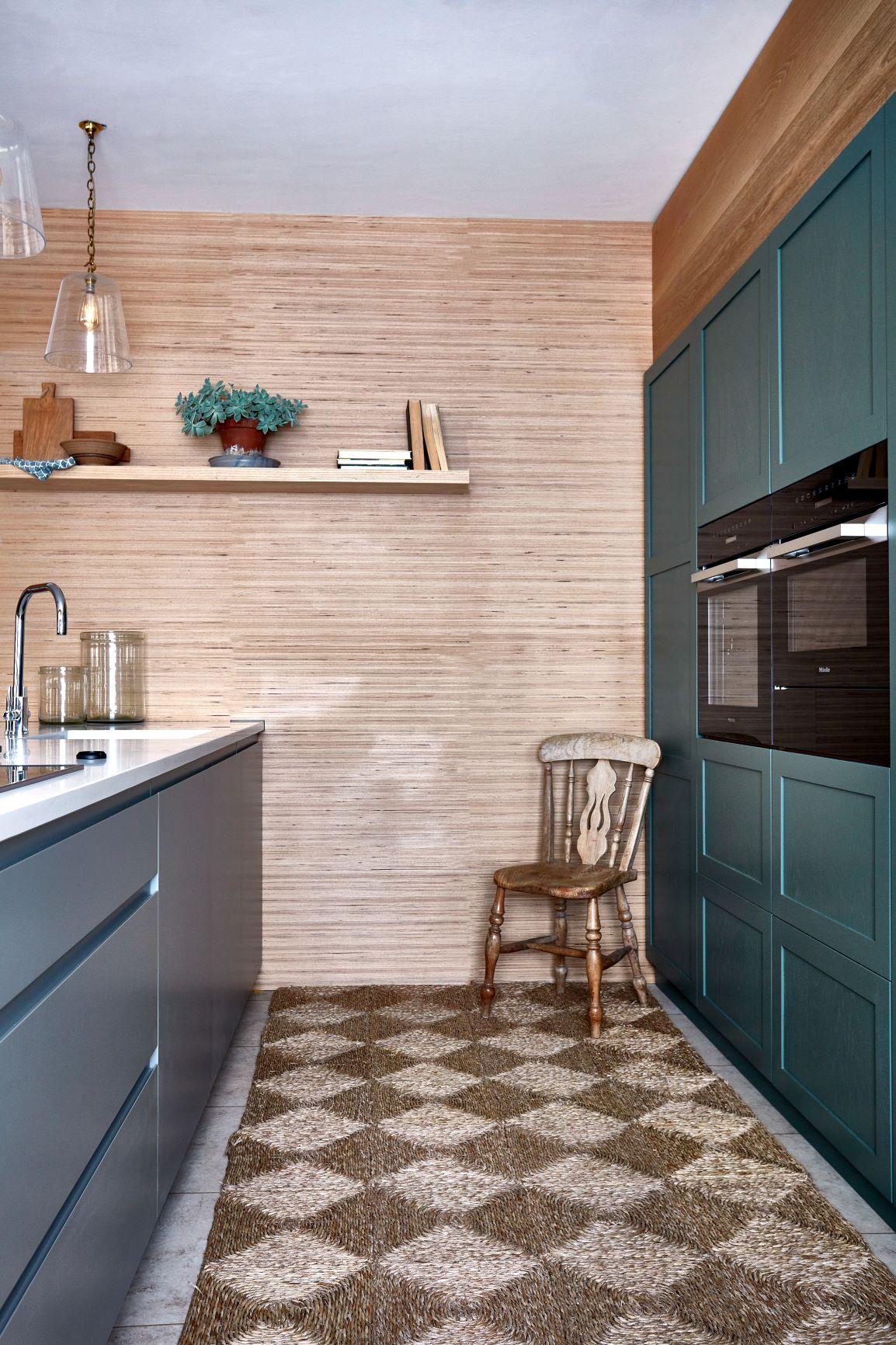 Modern kitchen with green cabinets and built-in oven, a wooden chair, and a geometric rug. Light wood paneling decorates the walls, with a white countertop and stainless steel faucet. A plant and books adorn the upper shelf. Pendant lights hang above.