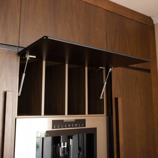 A modern kitchen cabinet with a lift-up door is open, revealing vertical wooden dividers for organized storage above a built-in appliance, all set in a dark wood finish.