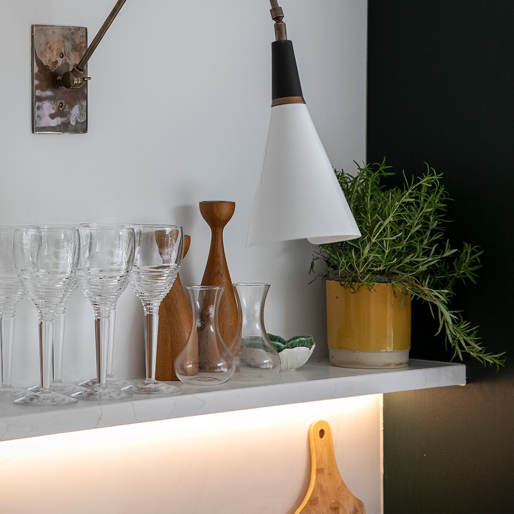 A white shelf holds glassware, a wooden vase, a small carafe, and a potted plant with green leaves. A modern white wall lamp hangs above, and a cutting board is partially visible below the shelf.