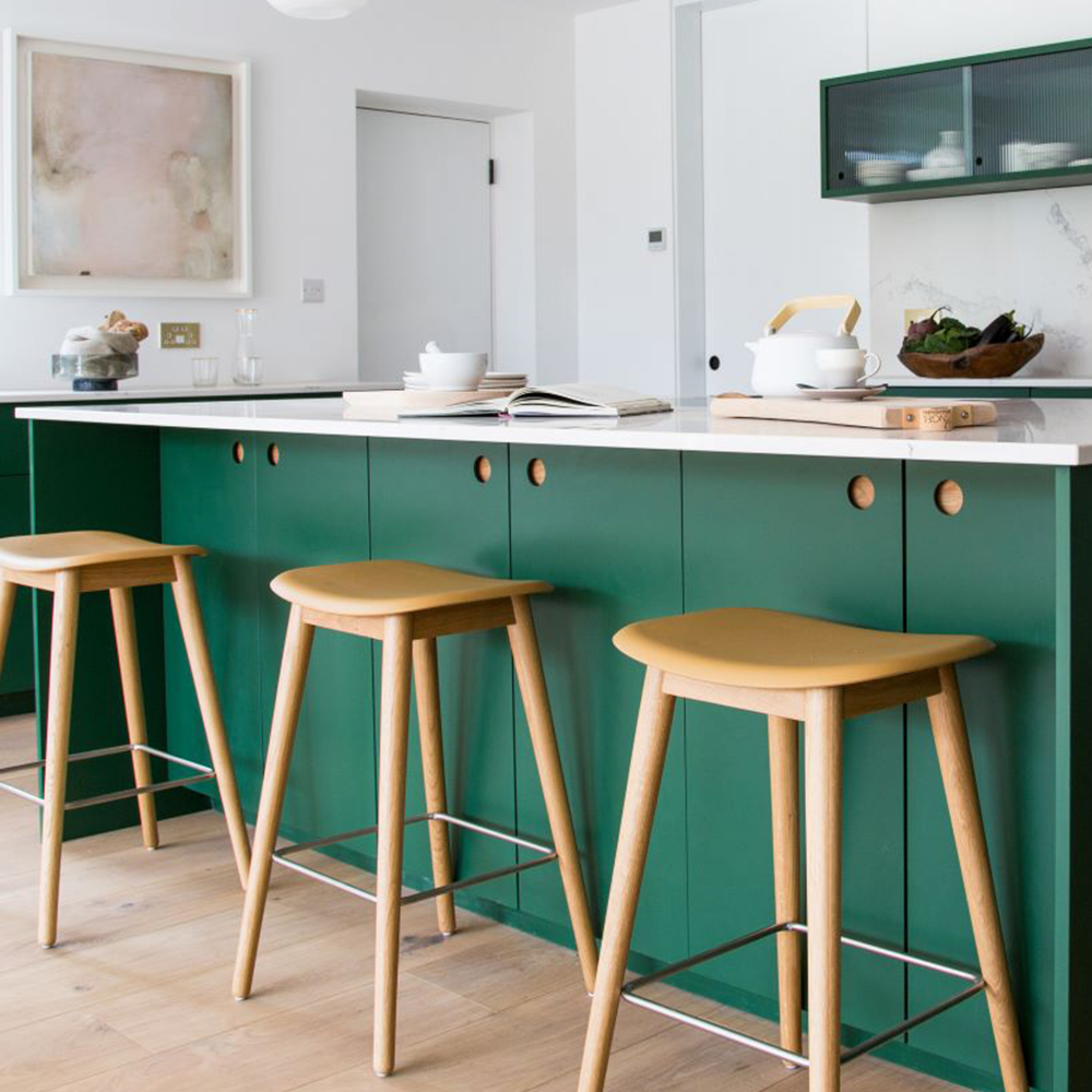 A modern kitchen with green cabinets, light wood bar stools, a white countertop, tea set, and decorative items. Light wood flooring and a minimal abstract painting complete the bright, airy space.