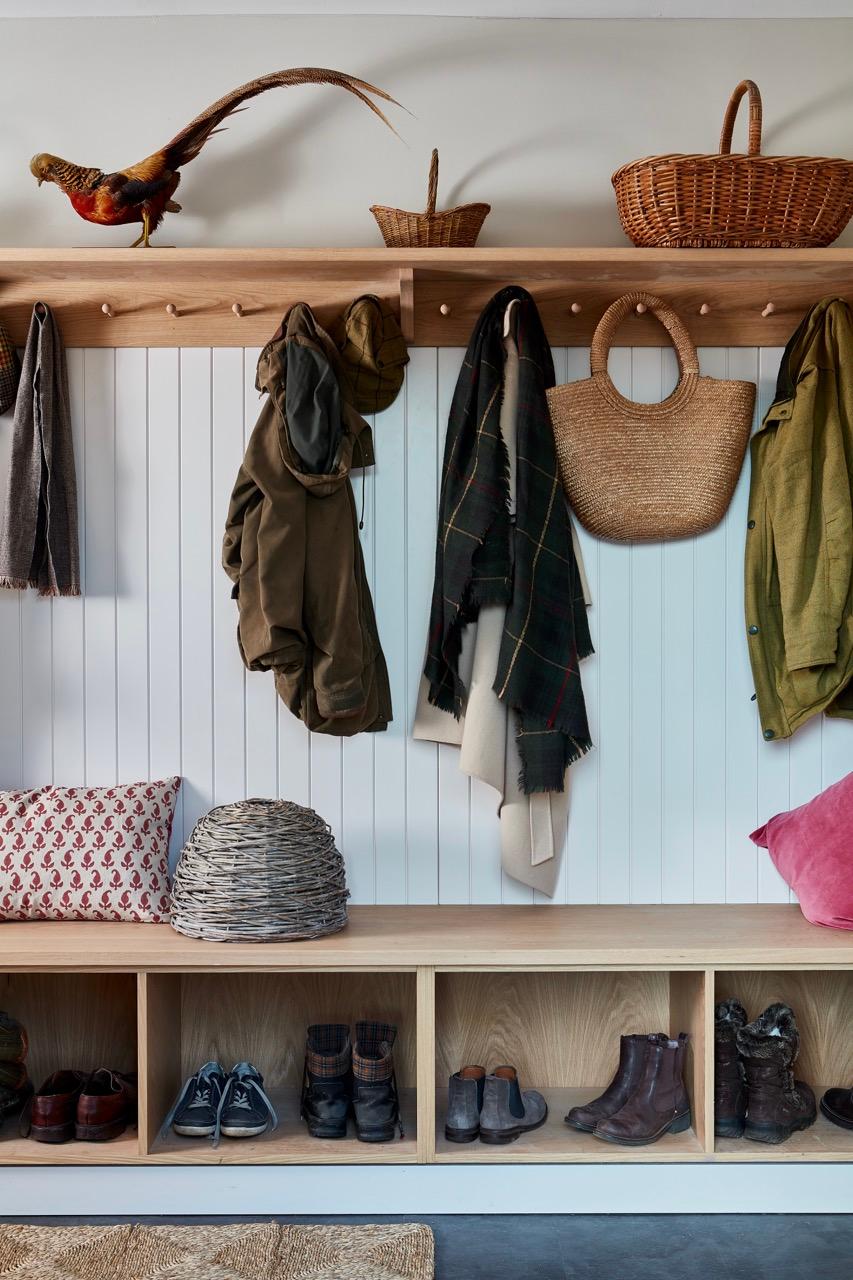 A cozy entryway features a wooden bench with cubby storage filled with shoes. Above, coats, scarves, and a woven bag hang on pegs. Decorative baskets and a pheasant figure sit on the wooden shelf above, creating a rustic and organized look.