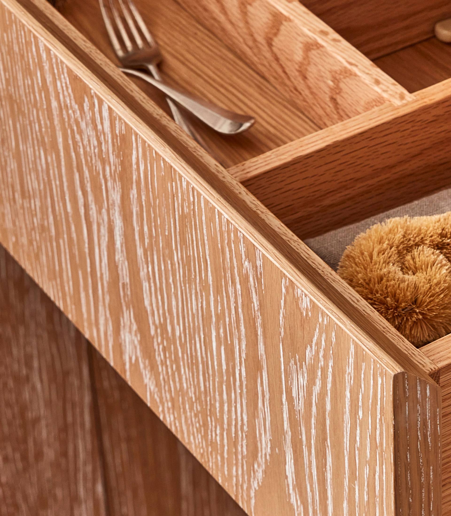 Open wooden drawer with a light oak finish, revealing a neatly organized interior. A fork is placed in one section, while a brown scrubber brush is visible in another compartment. The drawer has a textured, rustic look.