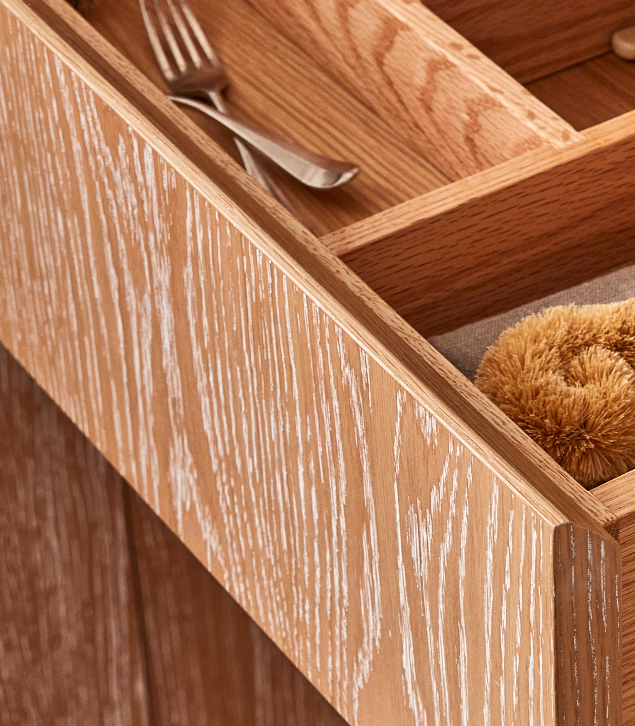 Open wooden drawer with a light oak finish, revealing a neatly organized interior. A fork is placed in one section, while a brown scrubber brush is visible in another compartment. The drawer has a textured, rustic look.