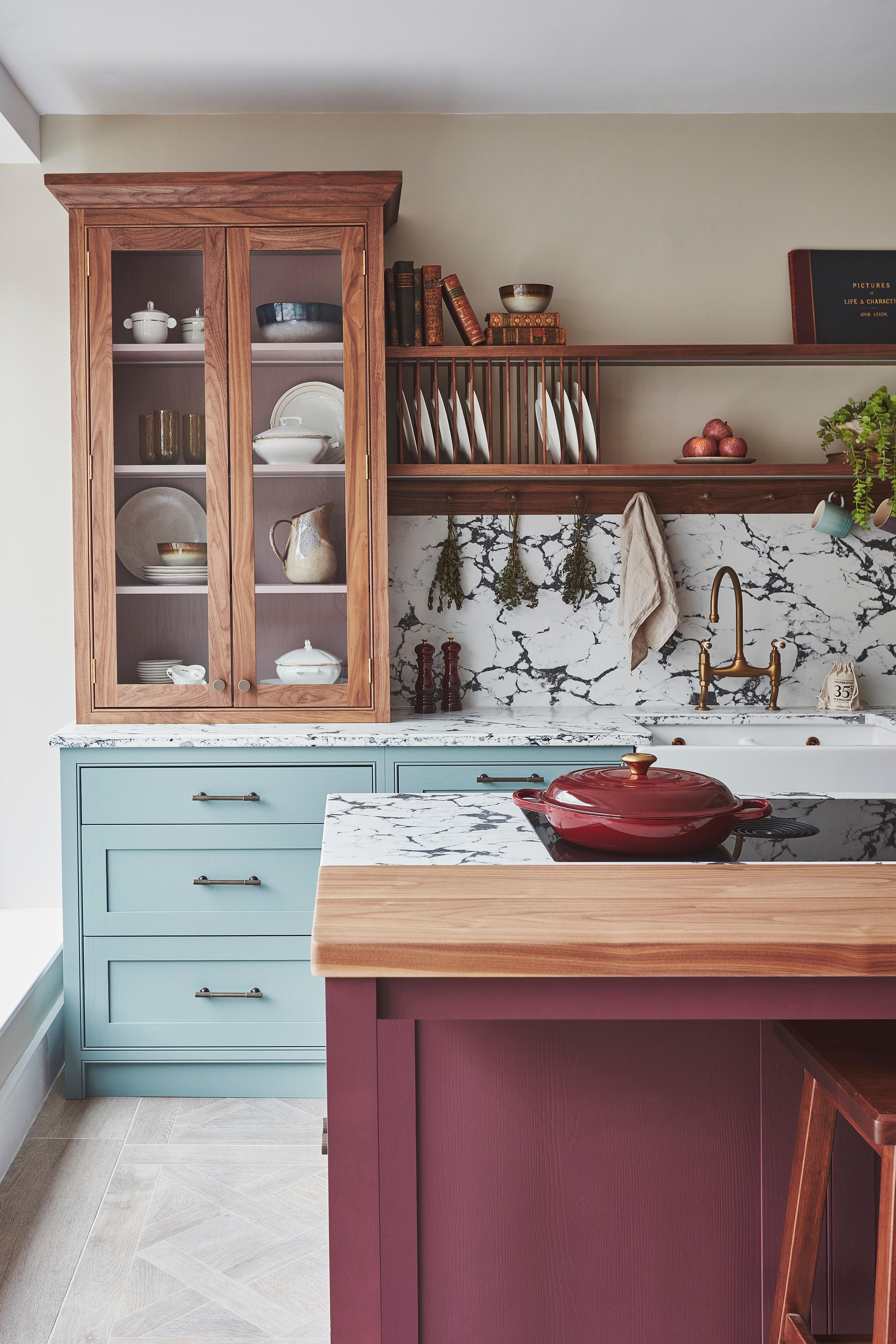 A cozy kitchen with a mix of styles. Light blue cabinets complement a marbled countertop and backsplash. A wooden shelf holds books and dishes, while a maroon island features a cutting board and red pot. Plants hang above the sink for decoration.