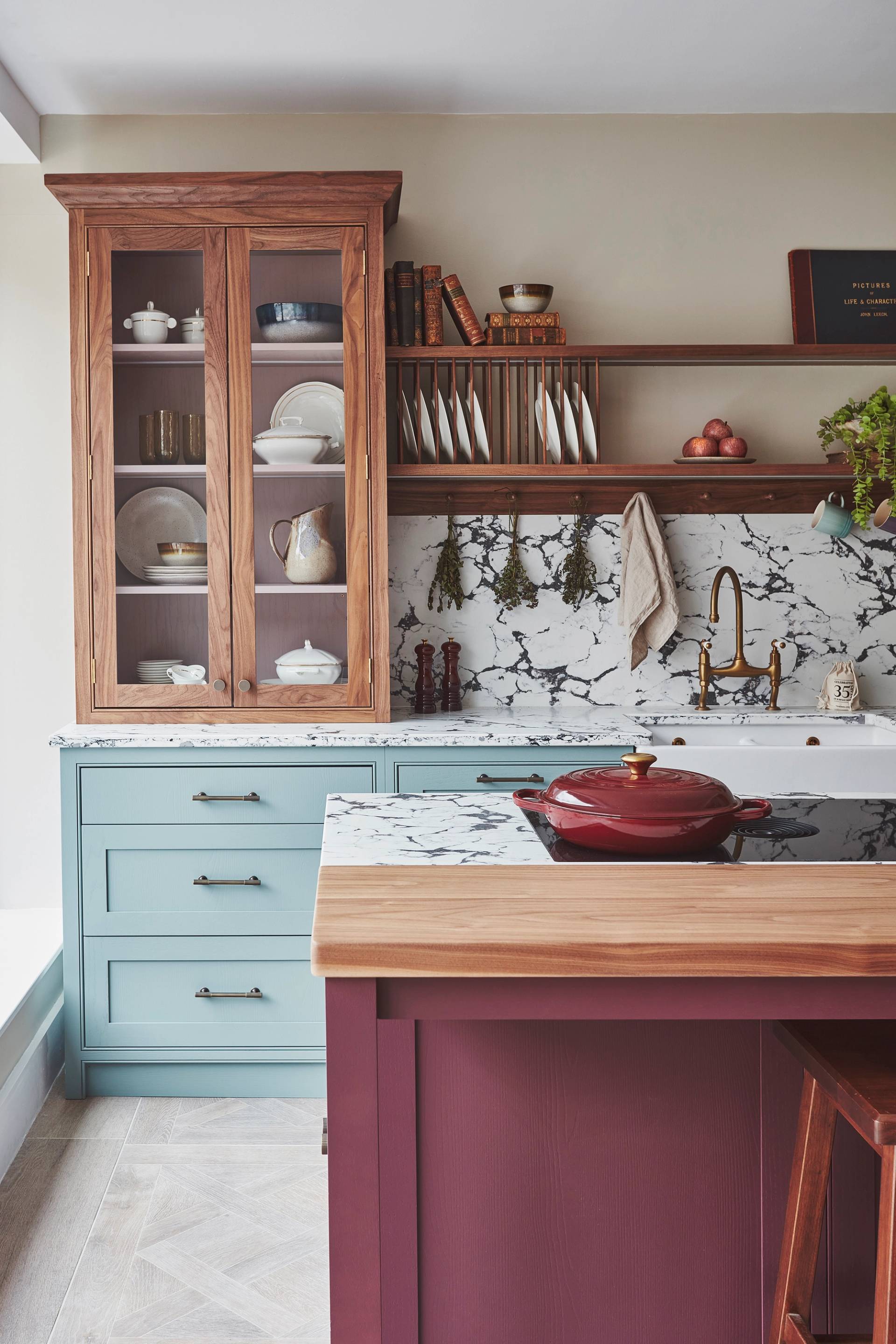 A cozy kitchen with a mix of styles. Light blue cabinets complement a marbled countertop and backsplash. A wooden shelf holds books and dishes, while a maroon island features a cutting board and red pot. Plants hang above the sink for decoration.