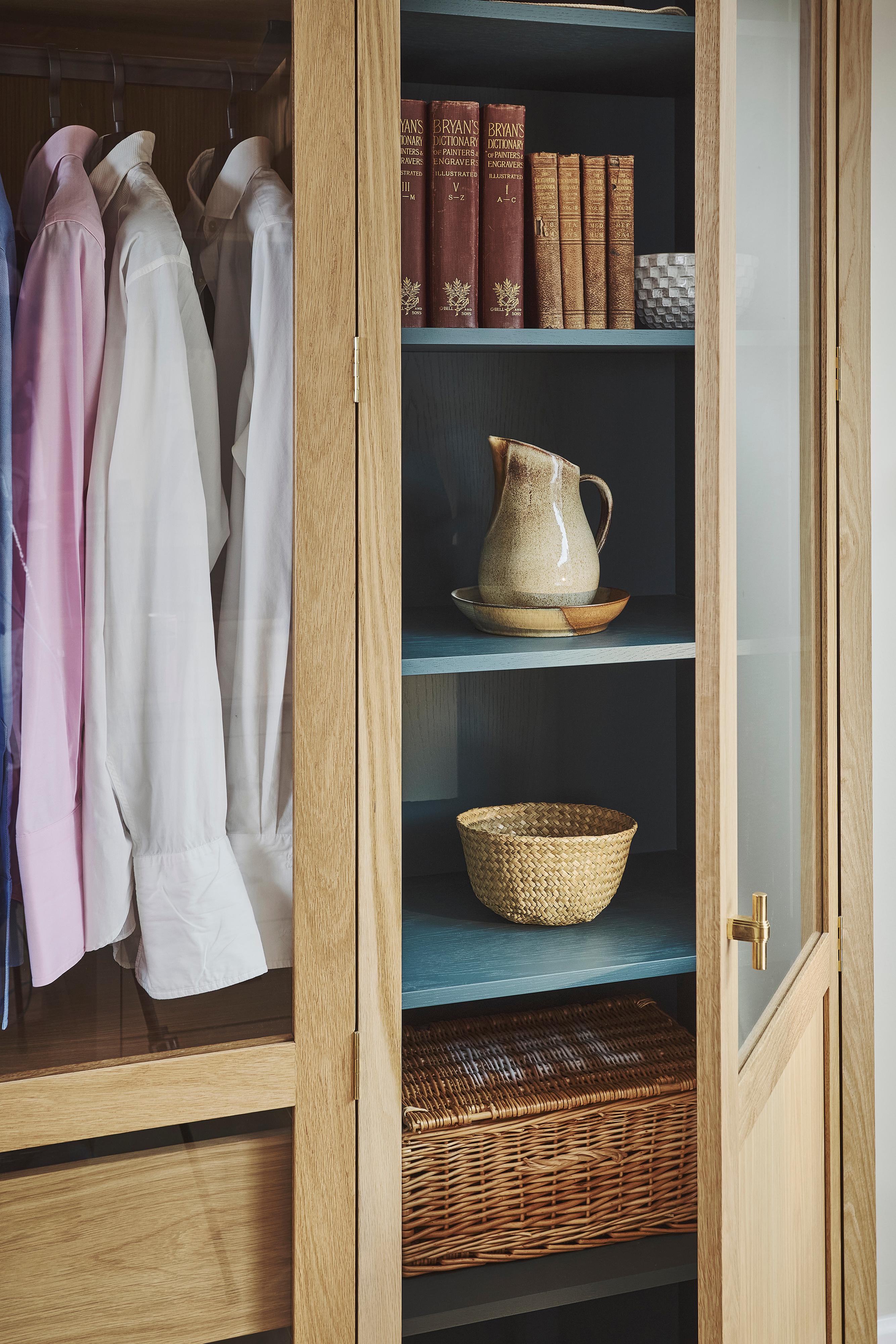 An oak wardrobe with glass doors displays neatly hung shirts. Adjacent open shelves hold books, a hat, a jug, a basket, and a wicker box, creating an organized and cozy storage space.