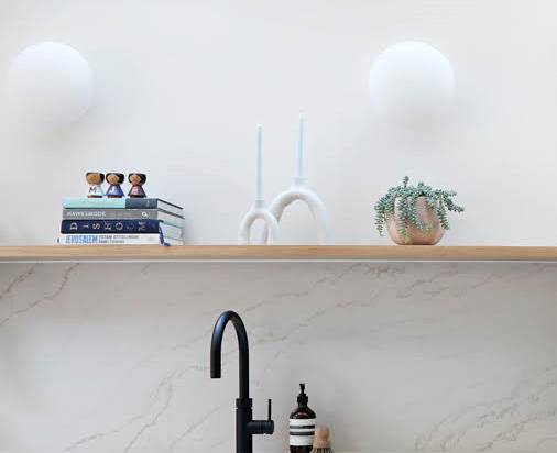 A wooden shelf above a marble countertop holds a small potted plant, two white candlesticks, stacked books, and small figurines. A black faucet and soap dispenser are visible below the shelf.