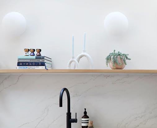 A wooden shelf above a marble countertop holds a small potted plant, two white candlesticks, stacked books, and small figurines. A black faucet and soap dispenser are visible below the shelf.