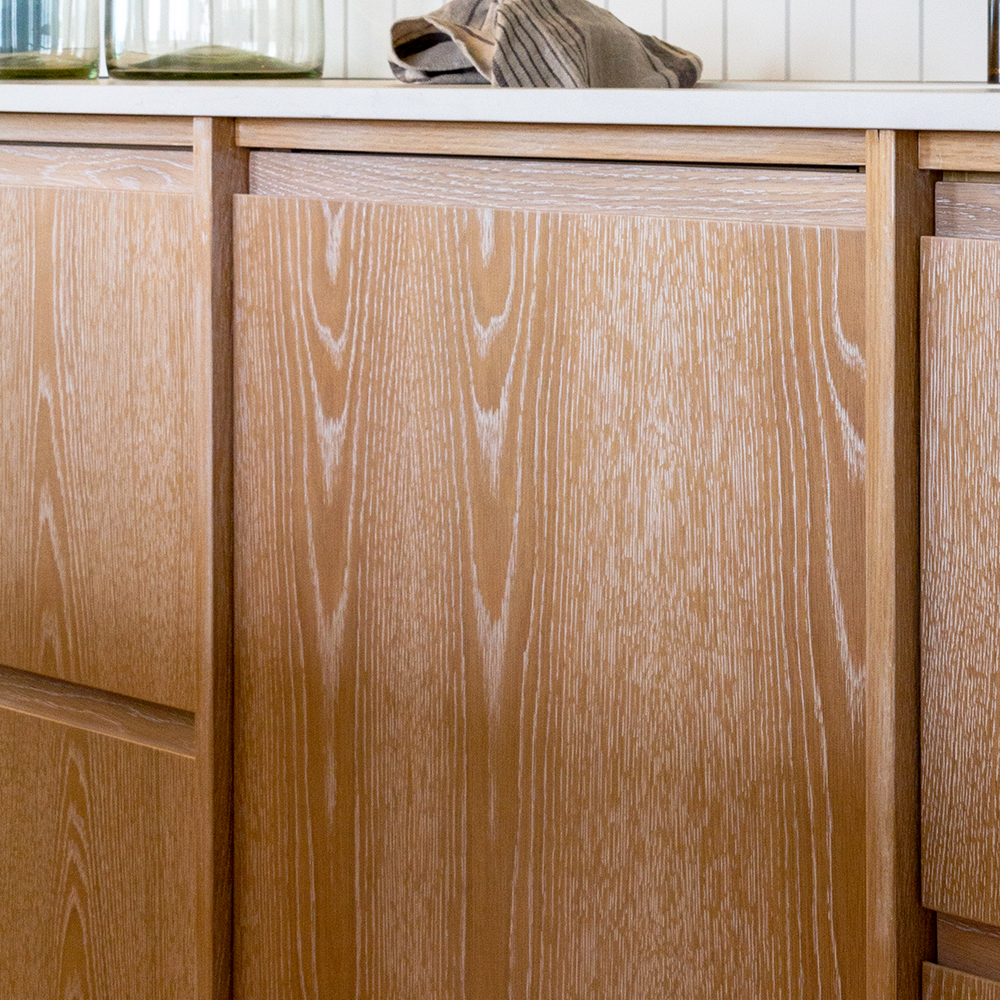 Cabinet with a light wood grain finish and sleek, handleless design. A white countertop is partially visible, supporting glass jars and a cloth.