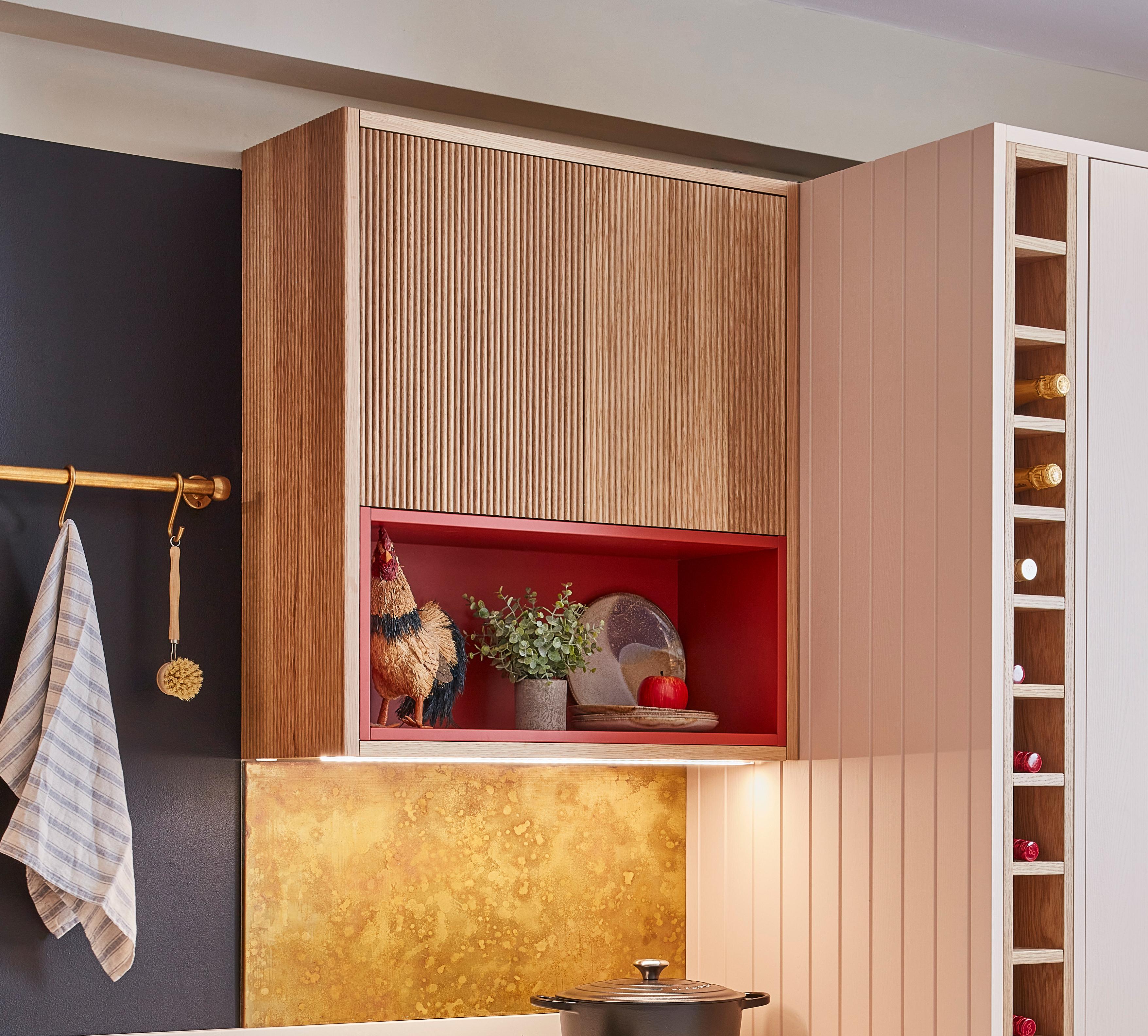 A modern kitchen corner with wooden cabinets, a red open shelf holding a rooster ornament, a plant, plates, and a red apple. A towel hangs on a rail, and a yellow-gold backsplash is above the stove.