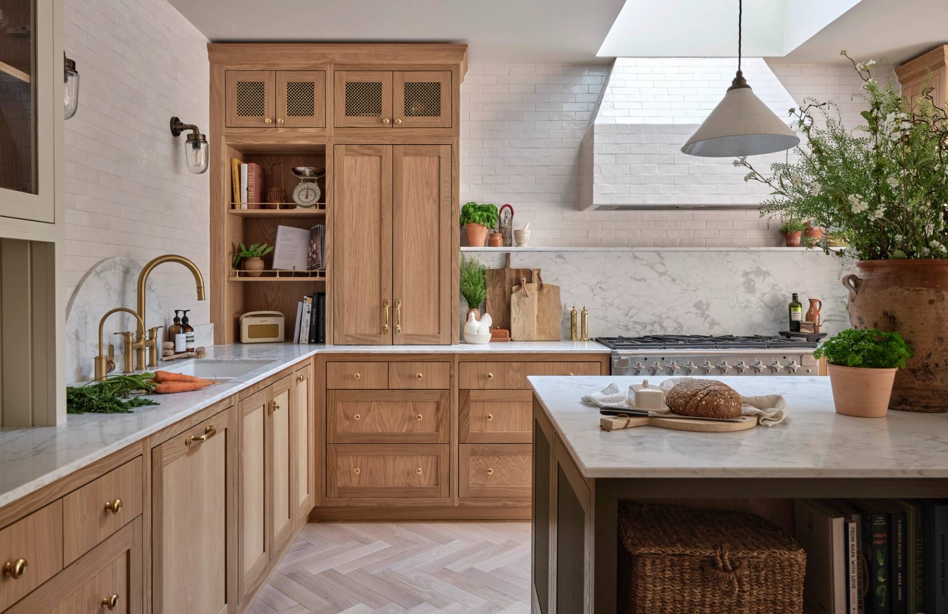 A bright, modern kitchen with light wood cabinets, marble countertops, brass fixtures, a gas stove, open shelves with decor, a kitchen island with fresh bread, and greenery in vases; natural light pours in from a skylight.