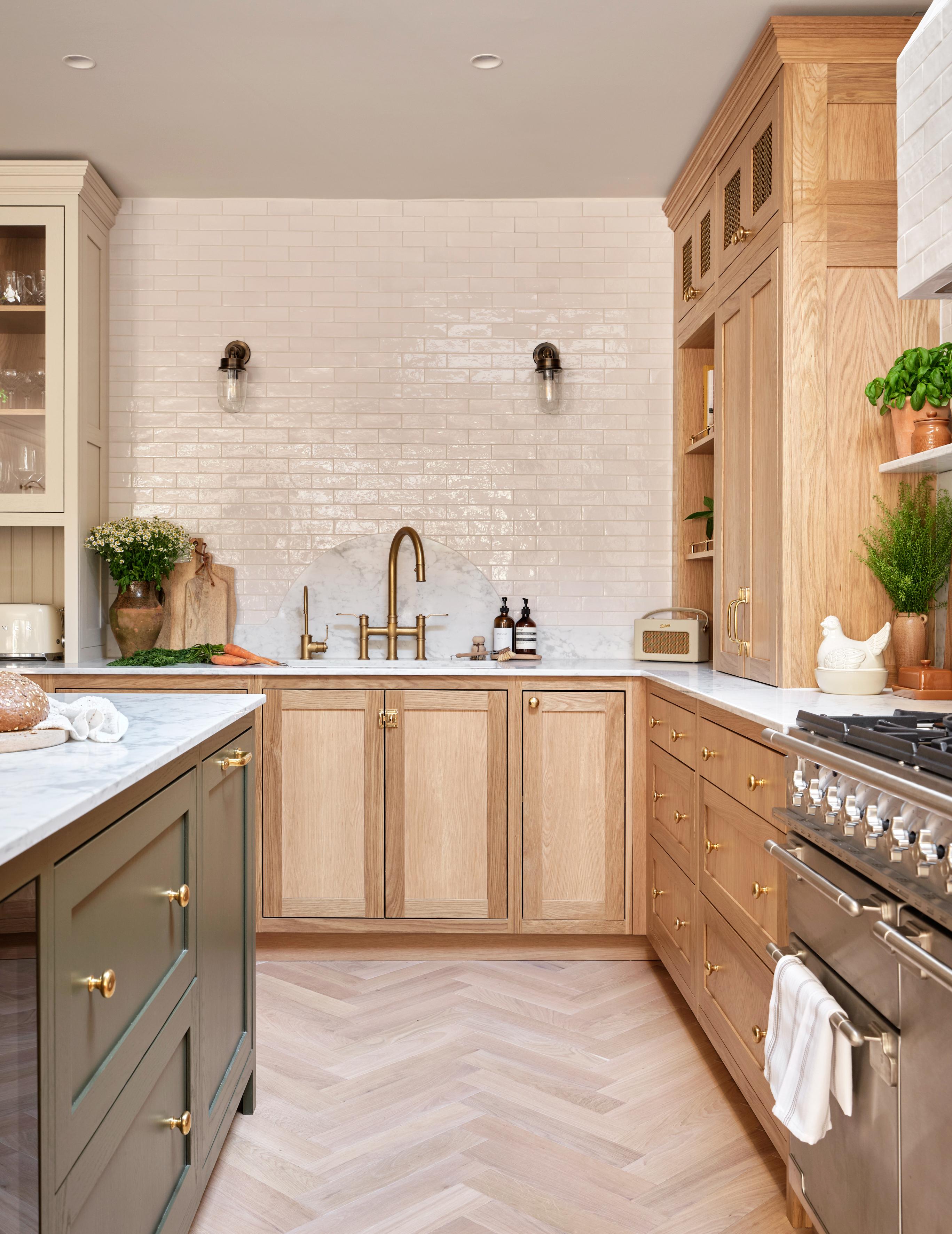 Modern kitchen with light wood cabinets, white subway tile backsplash, gold hardware, a farmhouse sink, marble countertops, stainless steel stove, and greenery in pots. Island has dark green cabinets and brass knobs.