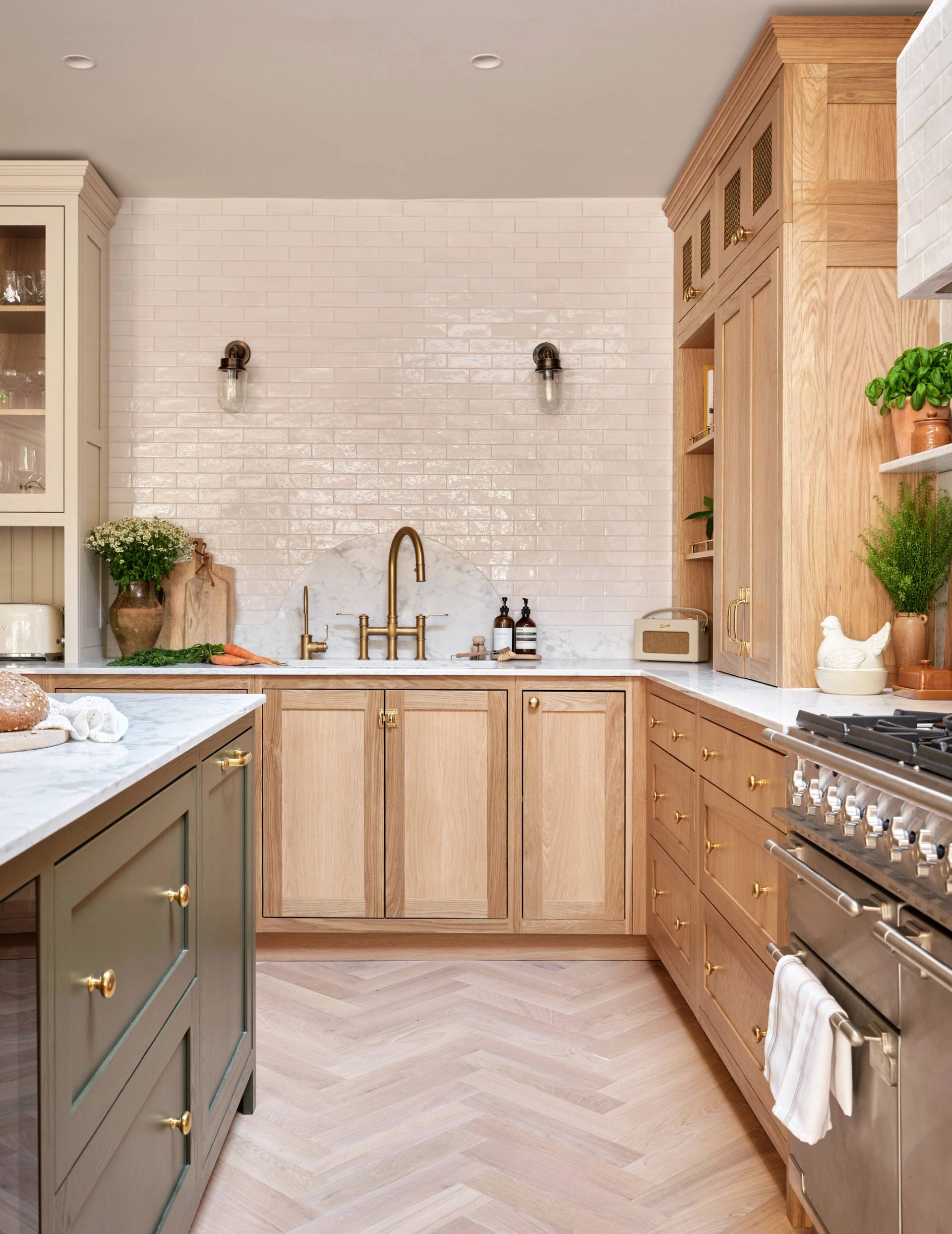 Modern kitchen with light wood cabinets, white subway tile backsplash, gold hardware, a farmhouse sink, marble countertops, stainless steel stove, and greenery in pots. Island has dark green cabinets and brass knobs.