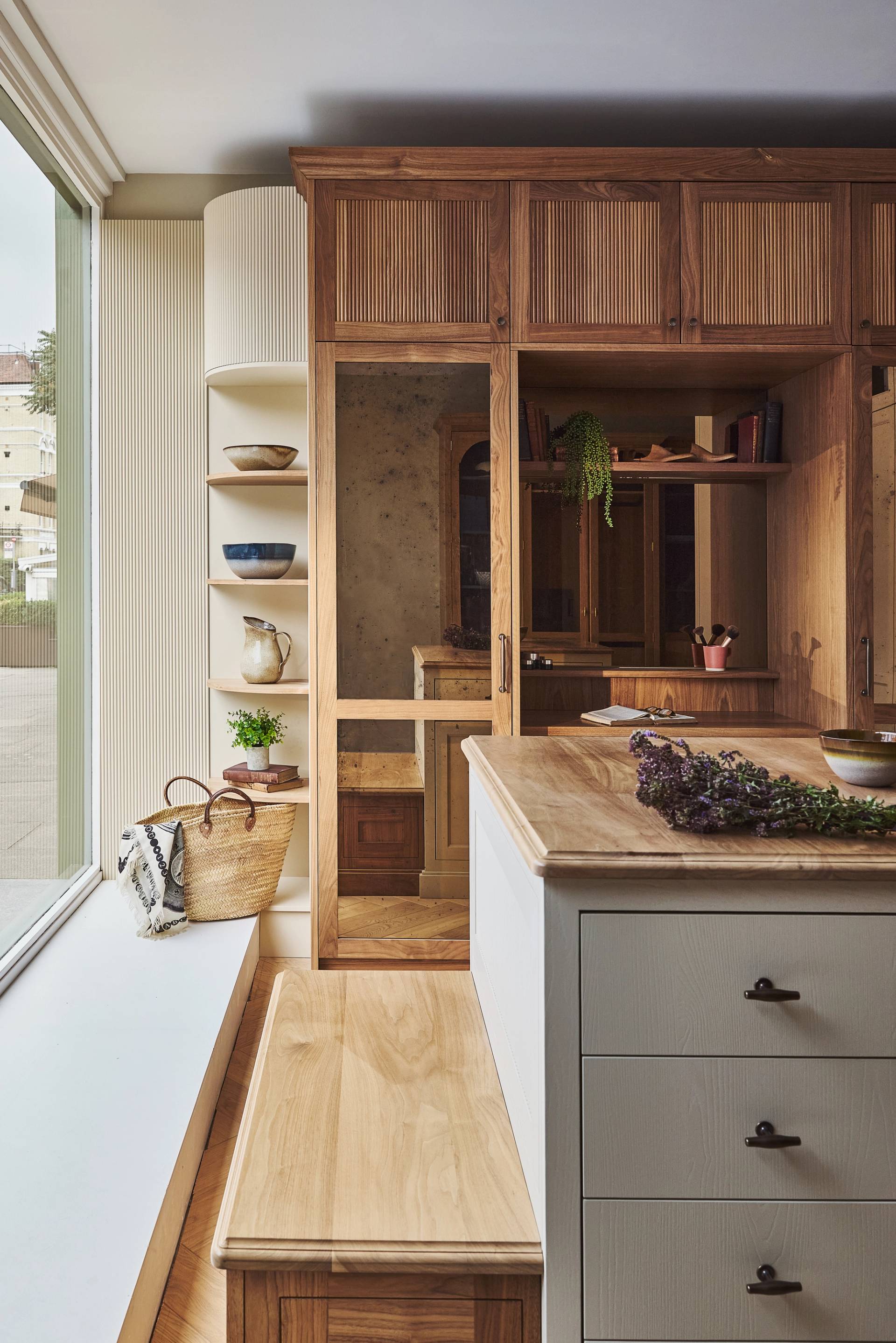 A modern kitchen with wooden cabinets, an open wardrobe displaying hanging clothes, open shelving with dishes, a kitchen island, and a large window letting in natural light. A basket and bench are by the window.
