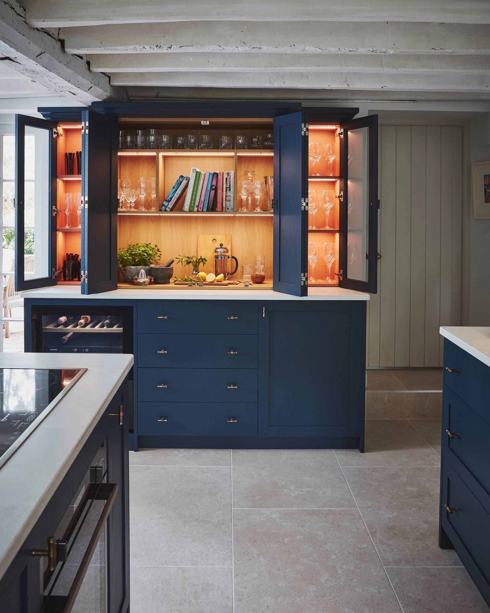 A kitchen cabinet with blue doors is open, revealing shelves with cookbooks, various glasses, a cutting board, herbs, a mortar and pestle, a glass pitcher, stacked glasses, and a halved lemon on a wooden counter.