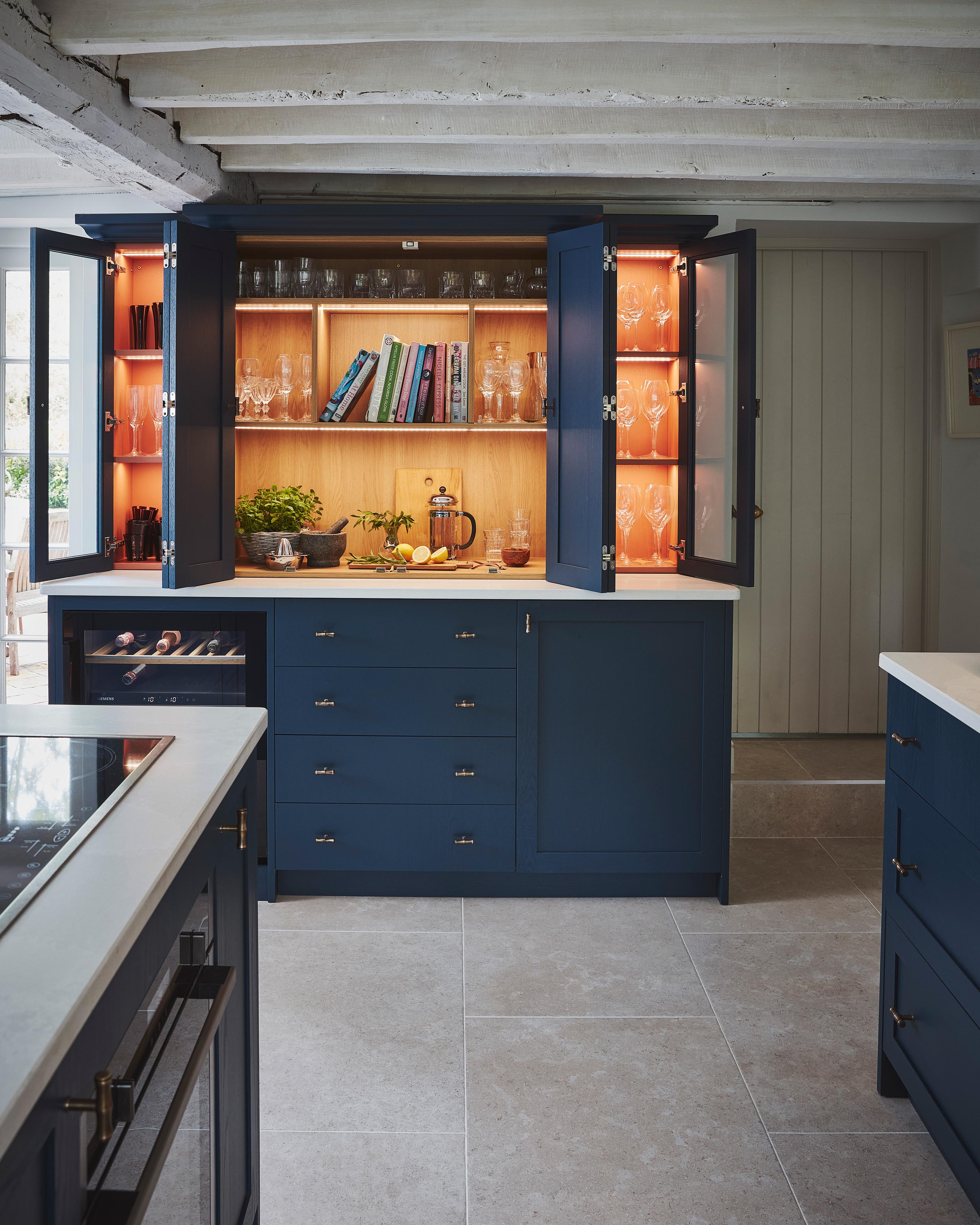 A kitchen cabinet with blue doors is open, revealing shelves with cookbooks, various glasses, a cutting board, herbs, a mortar and pestle, a glass pitcher, stacked glasses, and a halved lemon on a wooden counter.