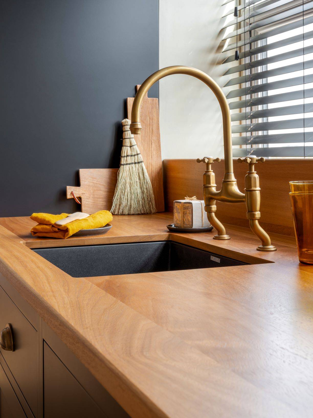 A modern kitchen counter with a wooden finish, featuring a brass faucet over a dark sink. A dish scrubber, an orange towel, and a glass sit nearby. A window with horizontal blinds lets in natural light.