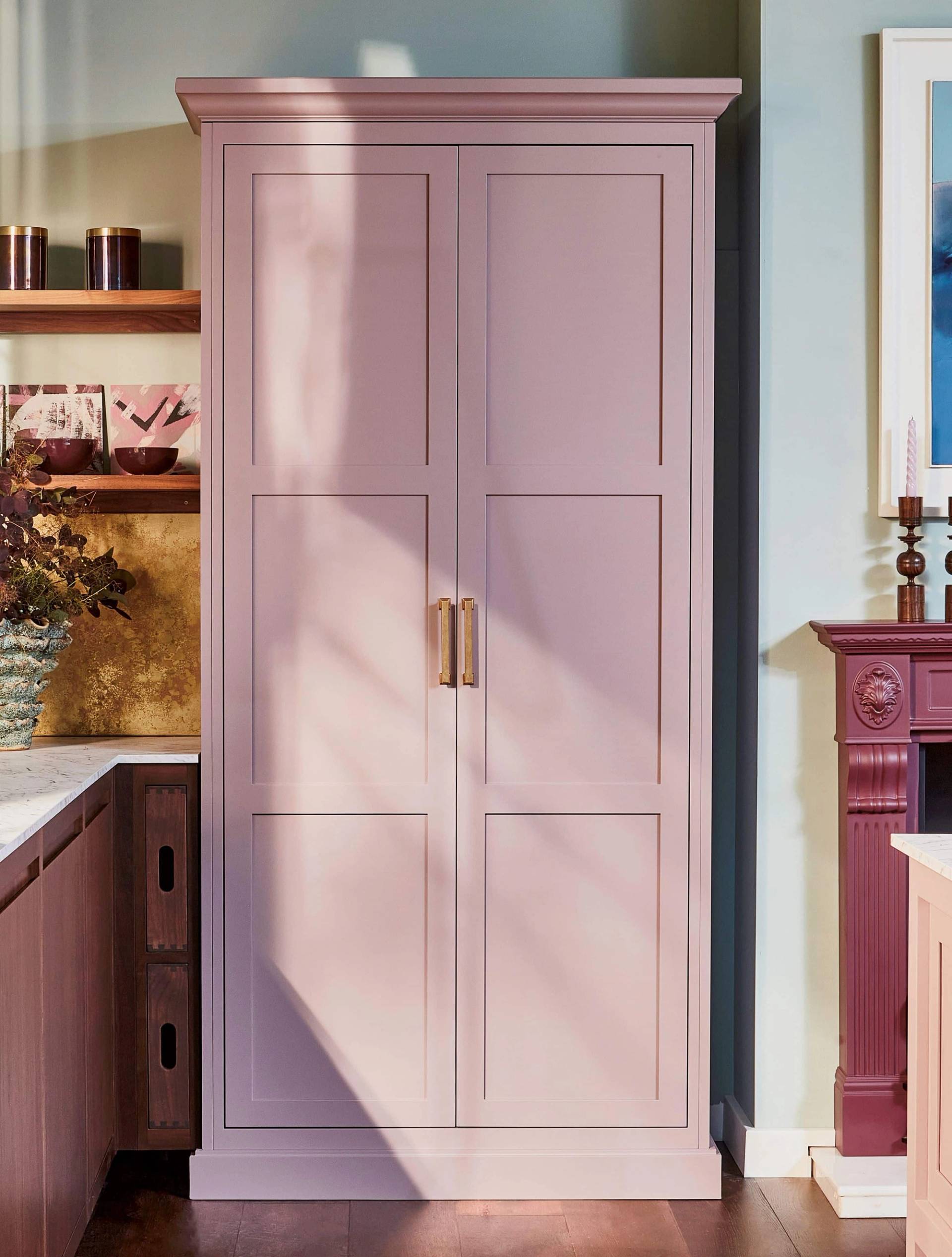 A tall, mauve cupboard with gold handles stands in a stylish kitchen next to a dark red fireplace, marble countertop, and decorative shelves with vases and jars. Sunlight casts soft shadows on the cupboard doors.