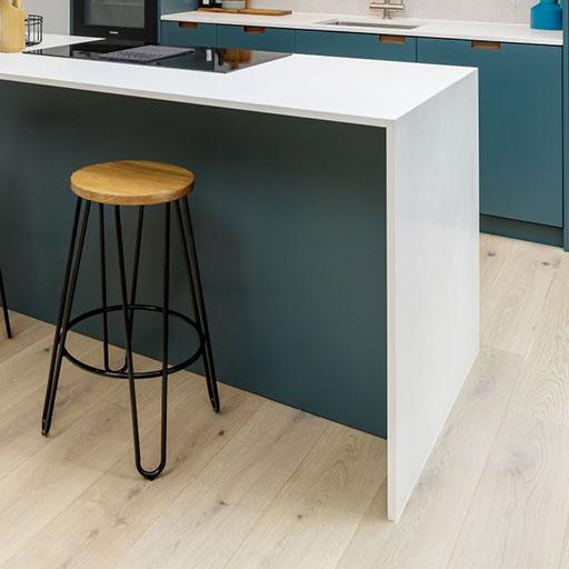 A modern kitchen with a white countertop island, a wooden stool with black metal legs, blue cabinets, and light wood flooring.