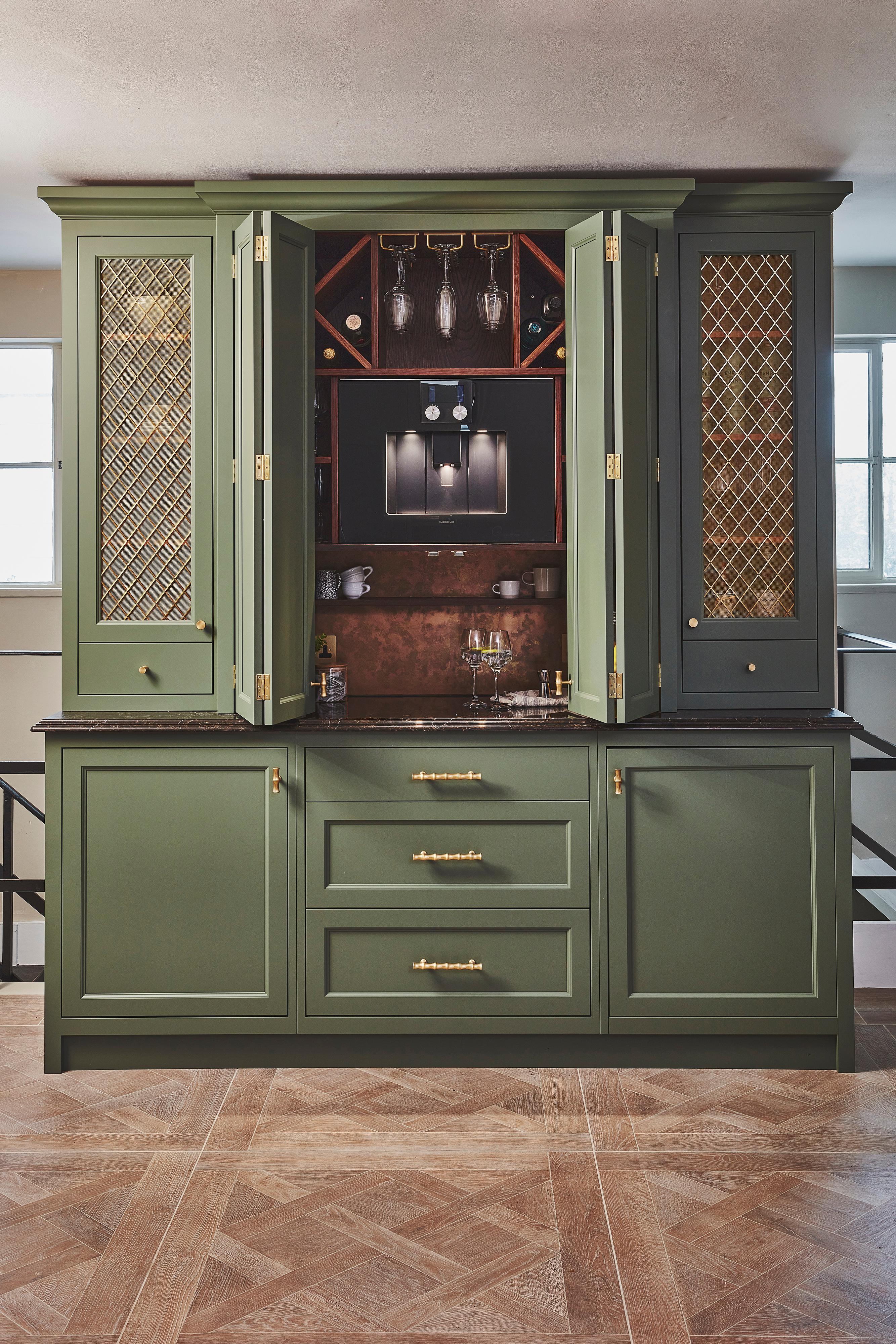 Sage green kitchen cabinets with gold mesh panel inserts, brass handles, and a dark countertop, set against a neutral wall with two windows in the background and geometric patterned wood flooring.