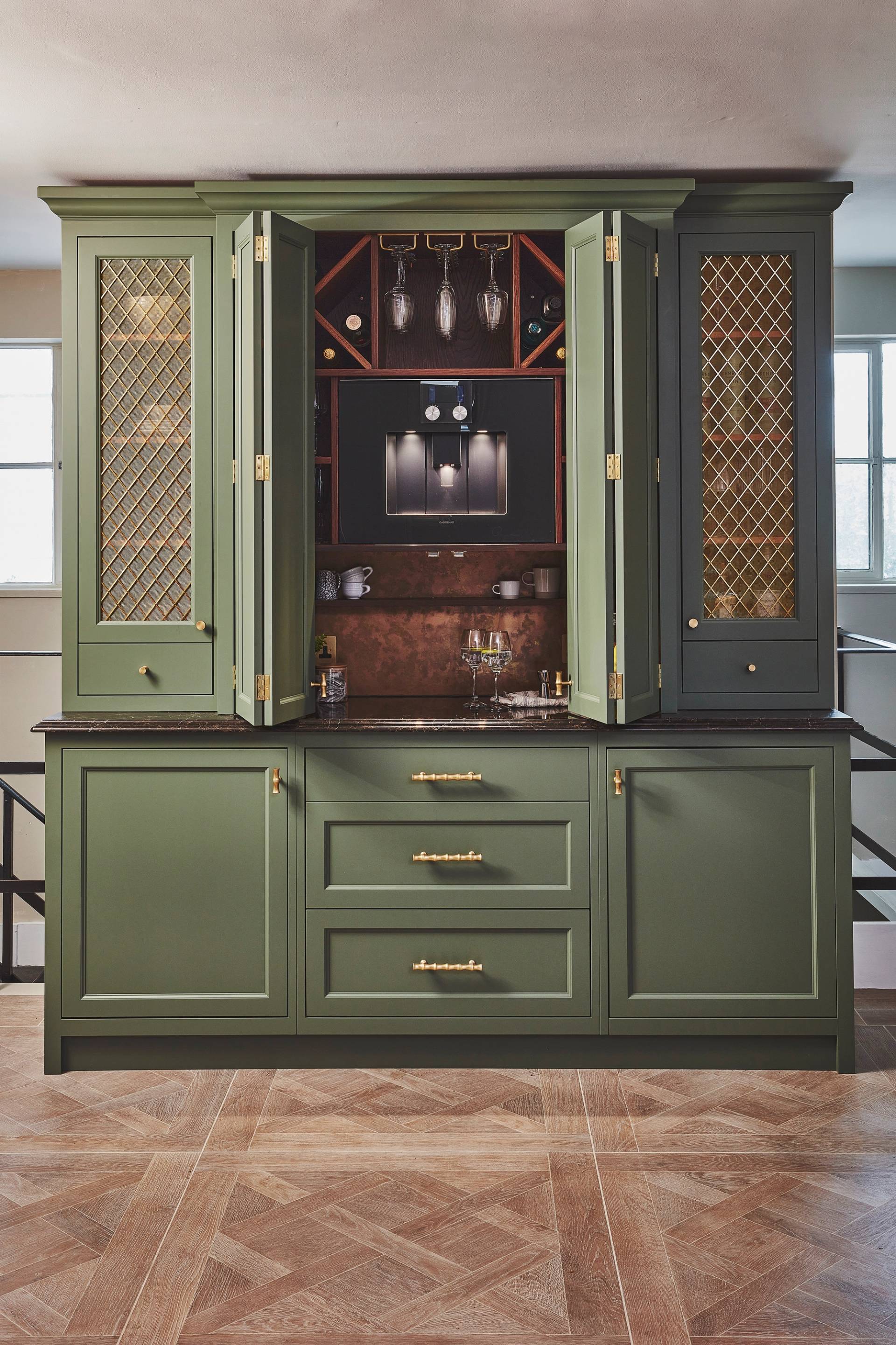 Sage green kitchen cabinets with gold mesh panel inserts, brass handles, and a dark countertop, set against a neutral wall with two windows in the background and geometric patterned wood flooring.