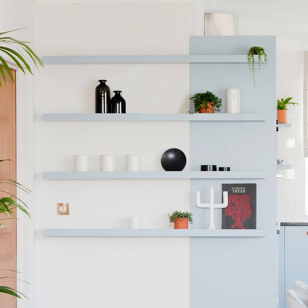 Four minimalist floating shelves on a two-tone wall hold plants, black and white vases, cups, a decorative candleholder, and a red book. The shelves display a modern, tidy arrangement.