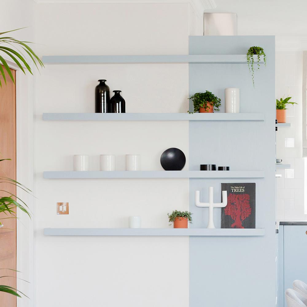 Four minimalist floating shelves on a two-tone wall hold plants, black and white vases, cups, a decorative candleholder, and a red book. The shelves display a modern, tidy arrangement.